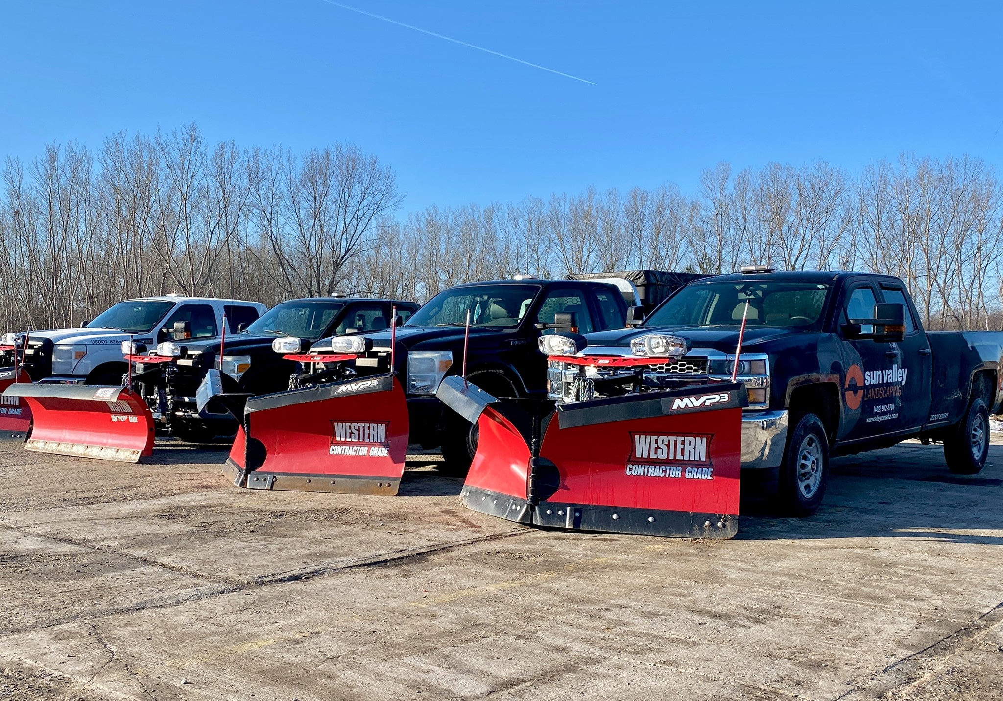 Three trucks equipped with red snow plows parked in a lot, set against a backdrop of leafless trees under a clear blue sky.