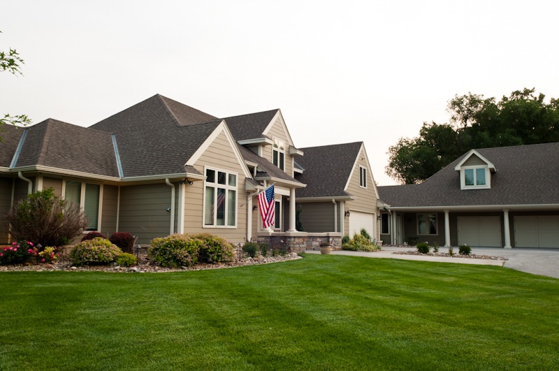 Spacious suburban house with a manicured lawn, American flag, driveway, and garage under a clear sky, surrounded by trees and shrubs.