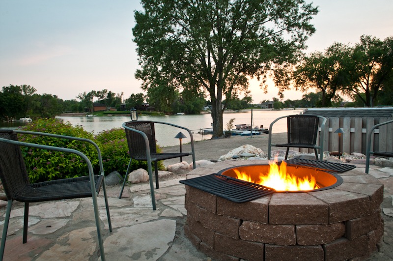 Cozy outdoor patio with fire pit, surrounded by trees and overlooking a peaceful lake at sunset. Empty chairs invite relaxation and warmth.