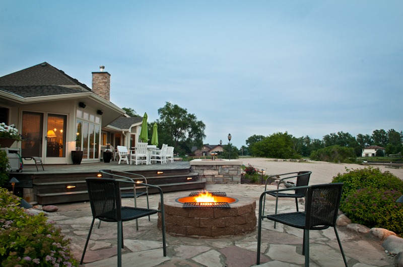 A cozy patio with a lit fire pit, surrounded by modern chairs and plants, adjacent to a house with large windows.