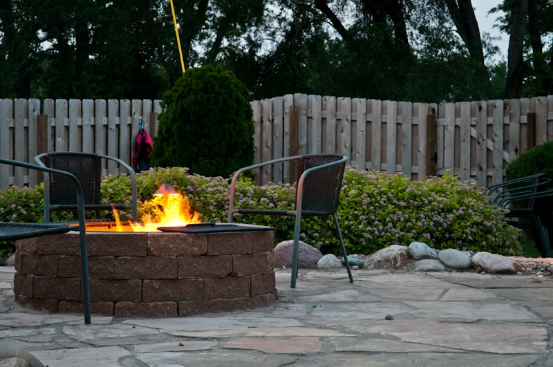 A backyard setting with a stone fire pit, surrounded by chairs and greenery, enclosed by a wooden fence under a dimly lit sky.