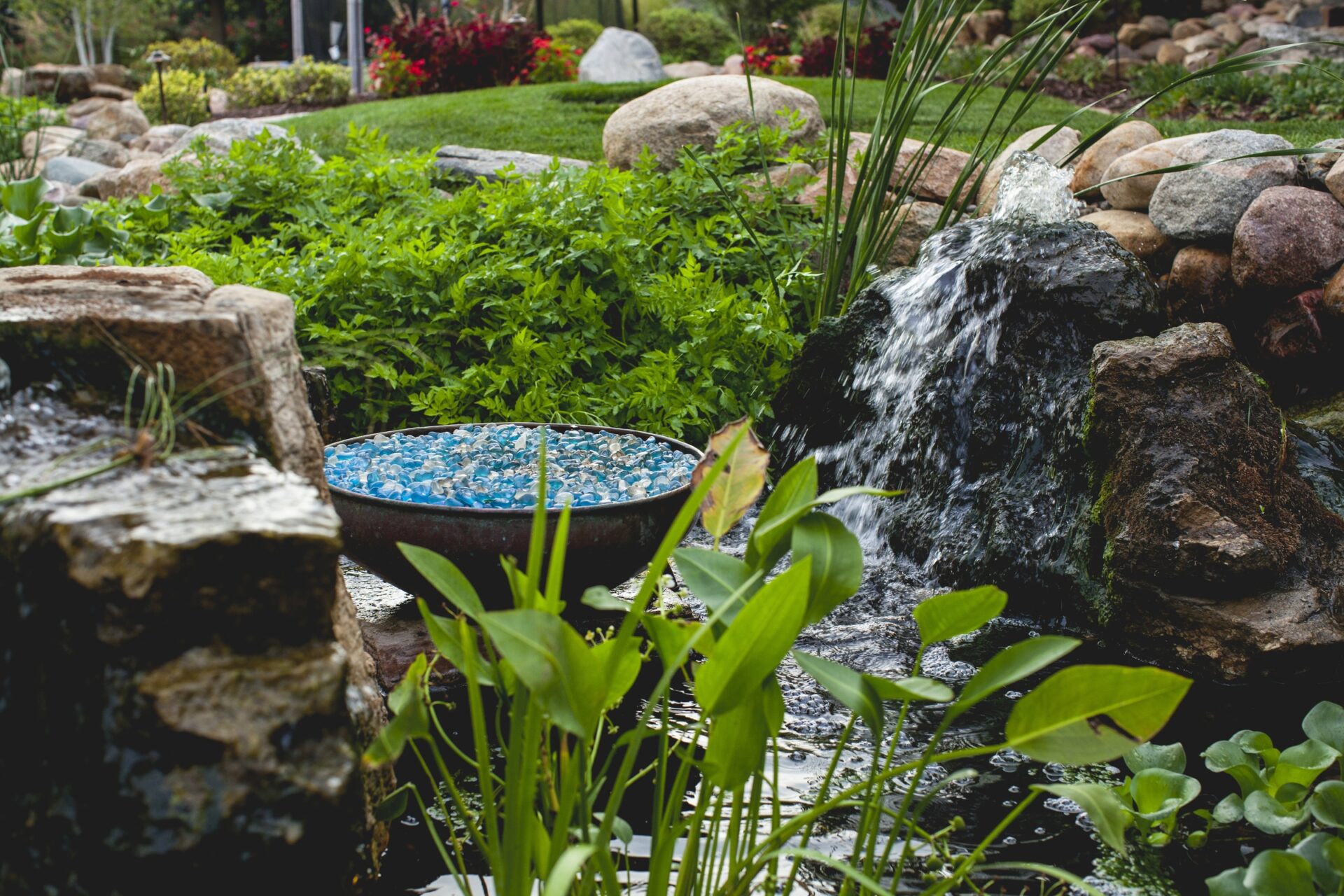 A serene garden with vibrant green foliage, a small waterfall flowing over rocks, and a dish filled with blue stones beside a pond.