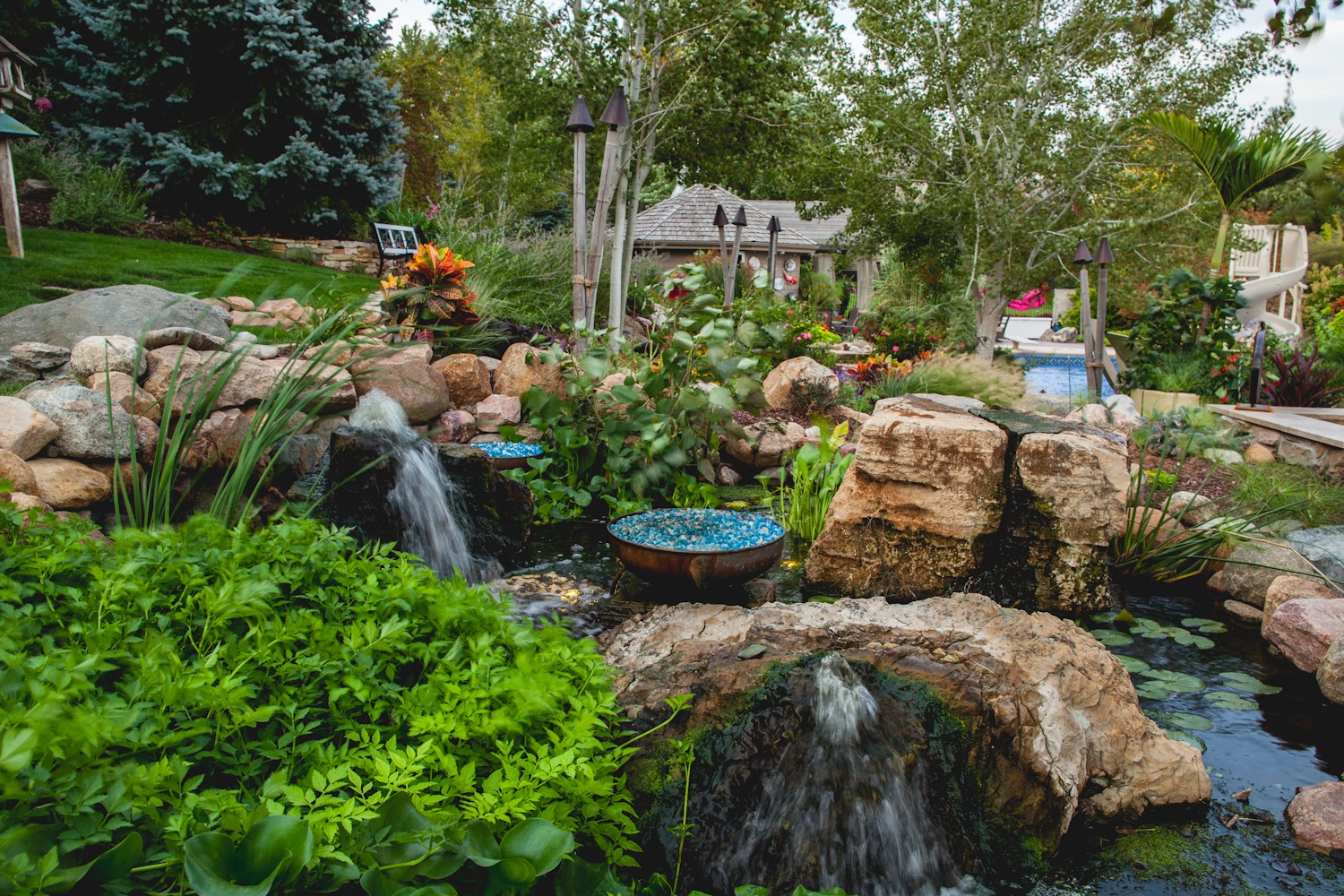 Lush garden with cascading waterfalls, vibrant plants, and blue glass art pieces create a serene, hidden oasis surrounded by trees and natural landscaping.