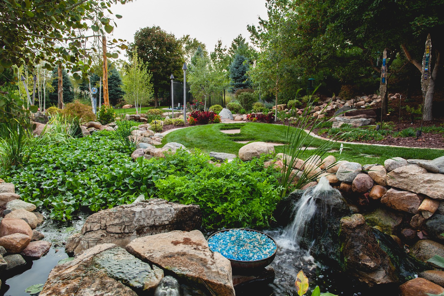 Lush garden with a small waterfall, colorful foliage, winding pathways, stones, and decorative tall sculptures surrounded by a variety of trees.