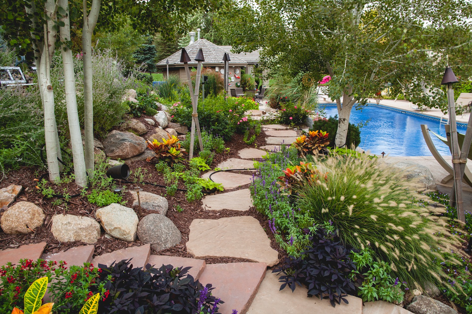 A serene garden path with lush greenery, colorful flowers, rocks, and a pool in the background. No people are present.