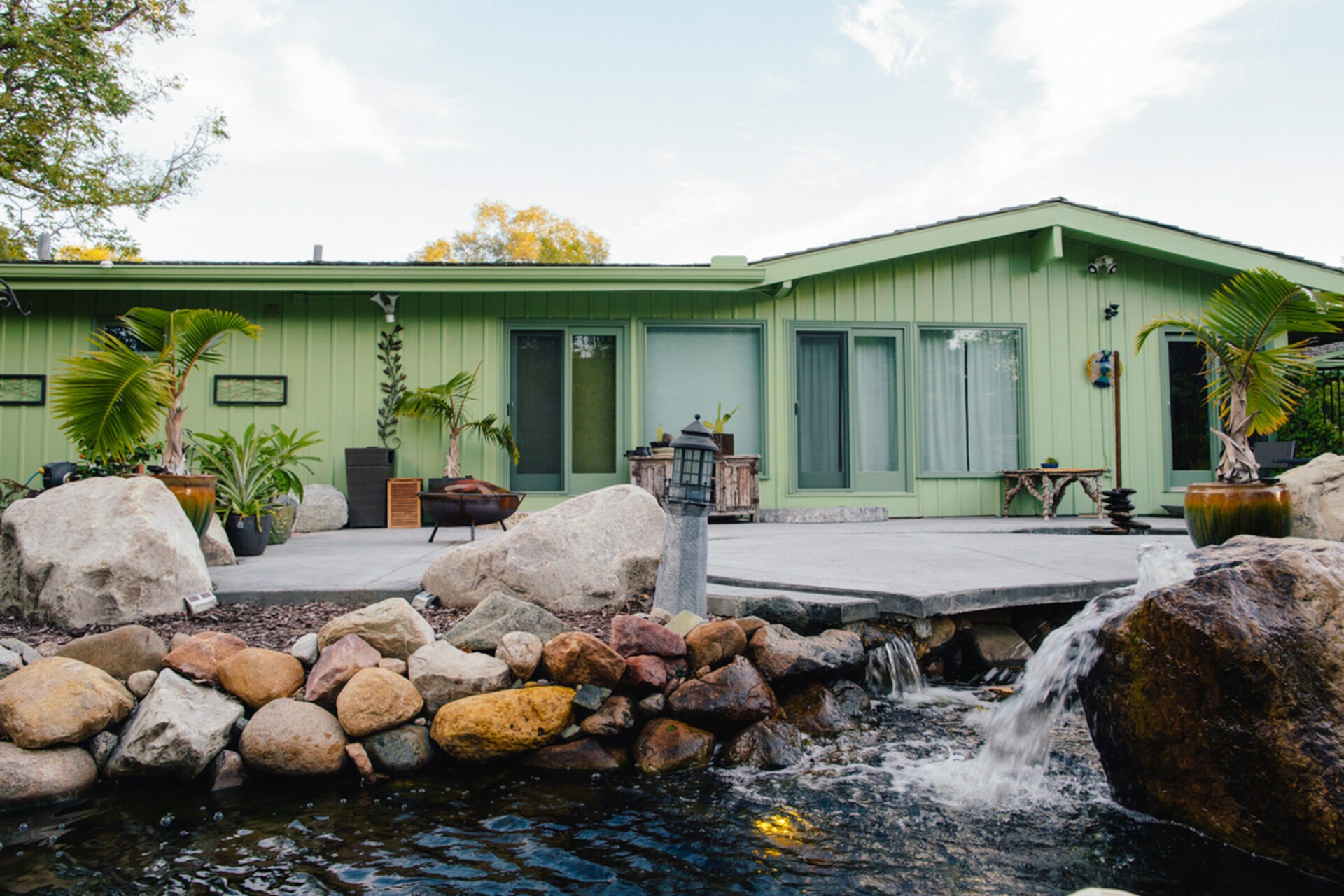 Single-story green house, surrounded by large rocks and lush plants, features a patio with a small pond and waterfall in the foreground.