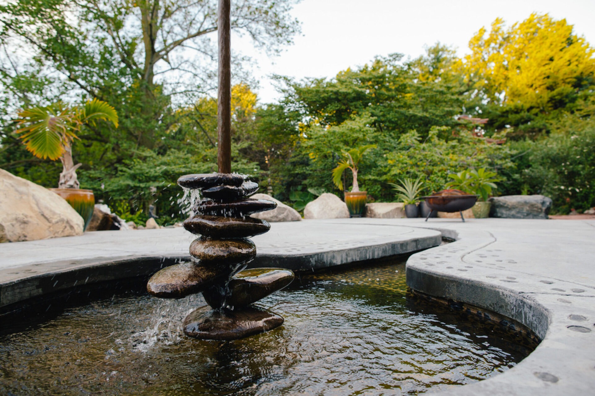 A serene garden features a unique stone fountain surrounded by lush greenery and decorative plants, creating a peaceful outdoor setting.