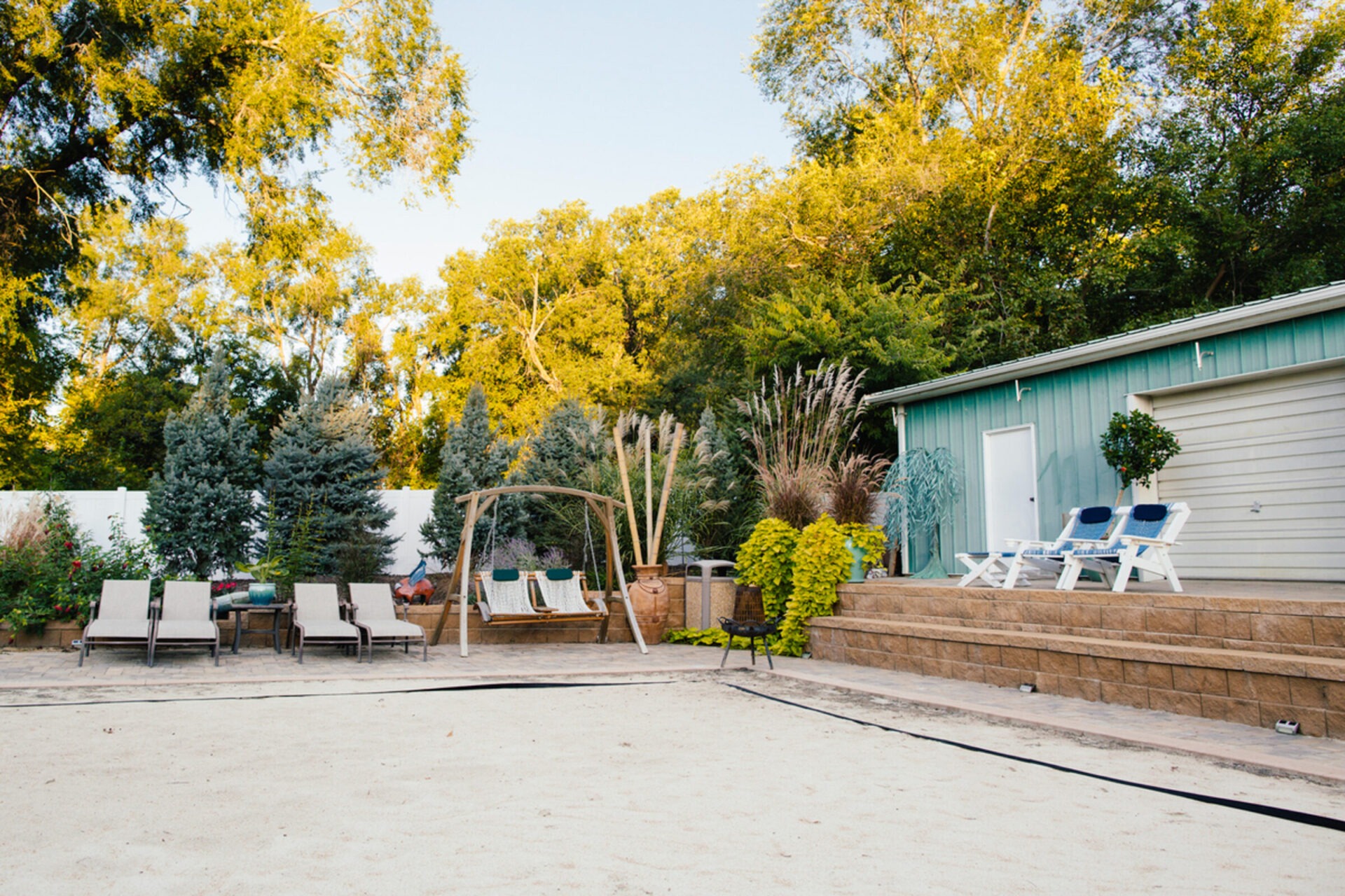 Backyard with loungers, swing, and plants. Trees and a small building with chairs on the raised patio. Peaceful, sunny setting.