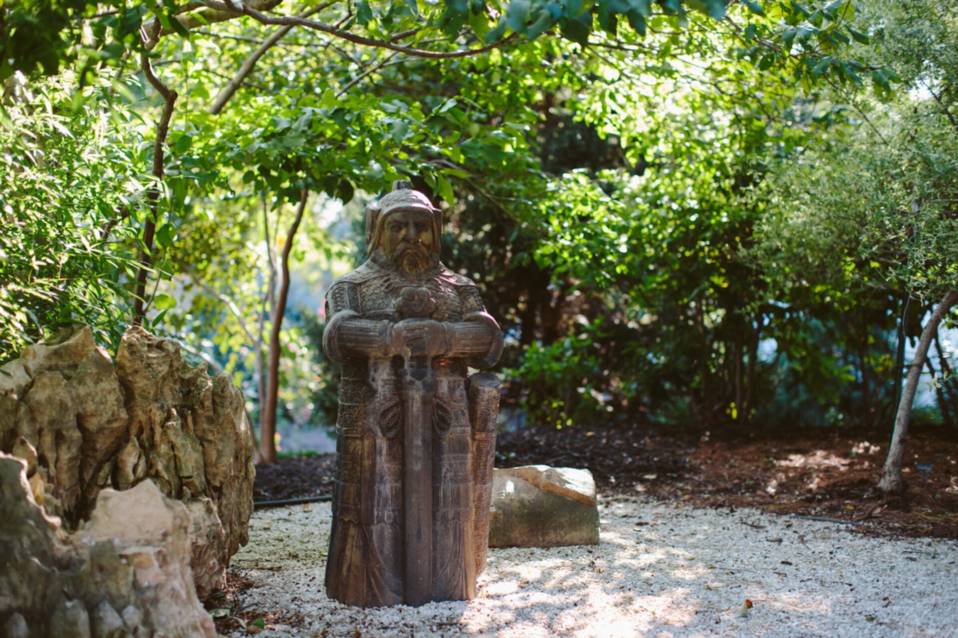 A stone statue of a person in armor stands in a lush garden, surrounded by trees and rocks, with dappled sunlight.