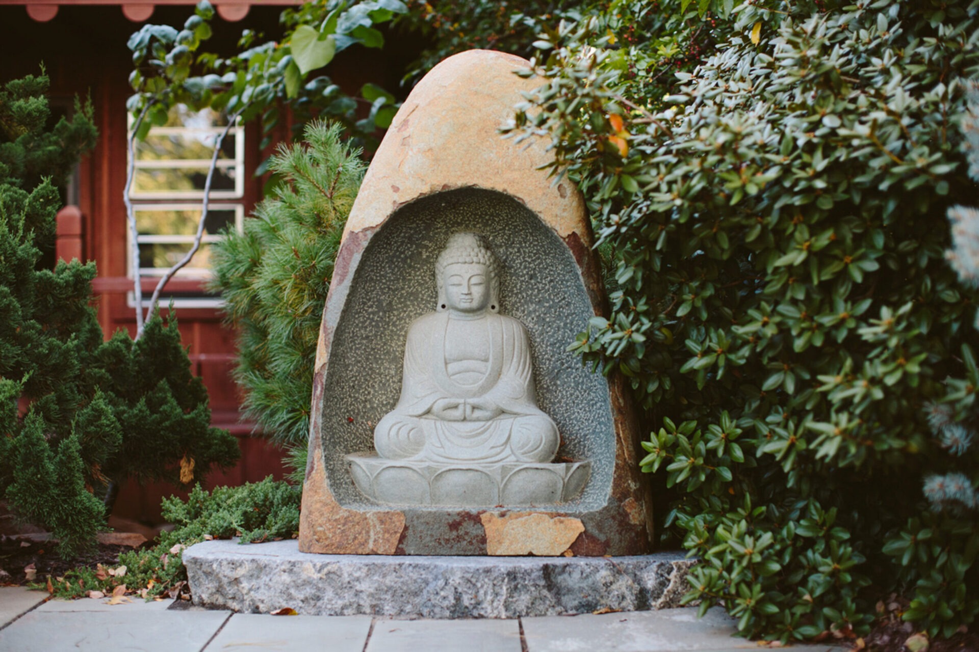 A stone Buddha statue is nestled in greenery near a wooden structure with window, creating a serene and tranquil atmosphere.