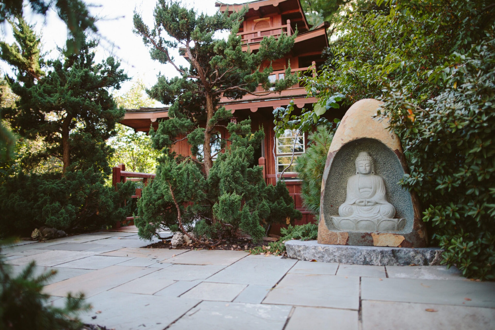 Serene garden featuring a Buddha statue set within stone, surrounded by trees and greenery, with a traditional wooden pavilion in the background.