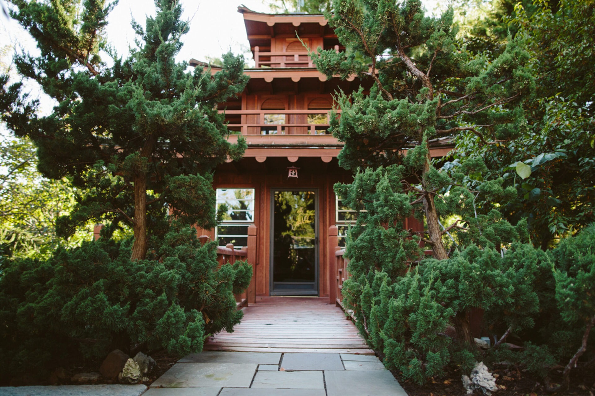 A traditional wooden Japanese-style teahouse surrounded by lush green trees and stone pathway, creating a serene, peaceful garden setting.