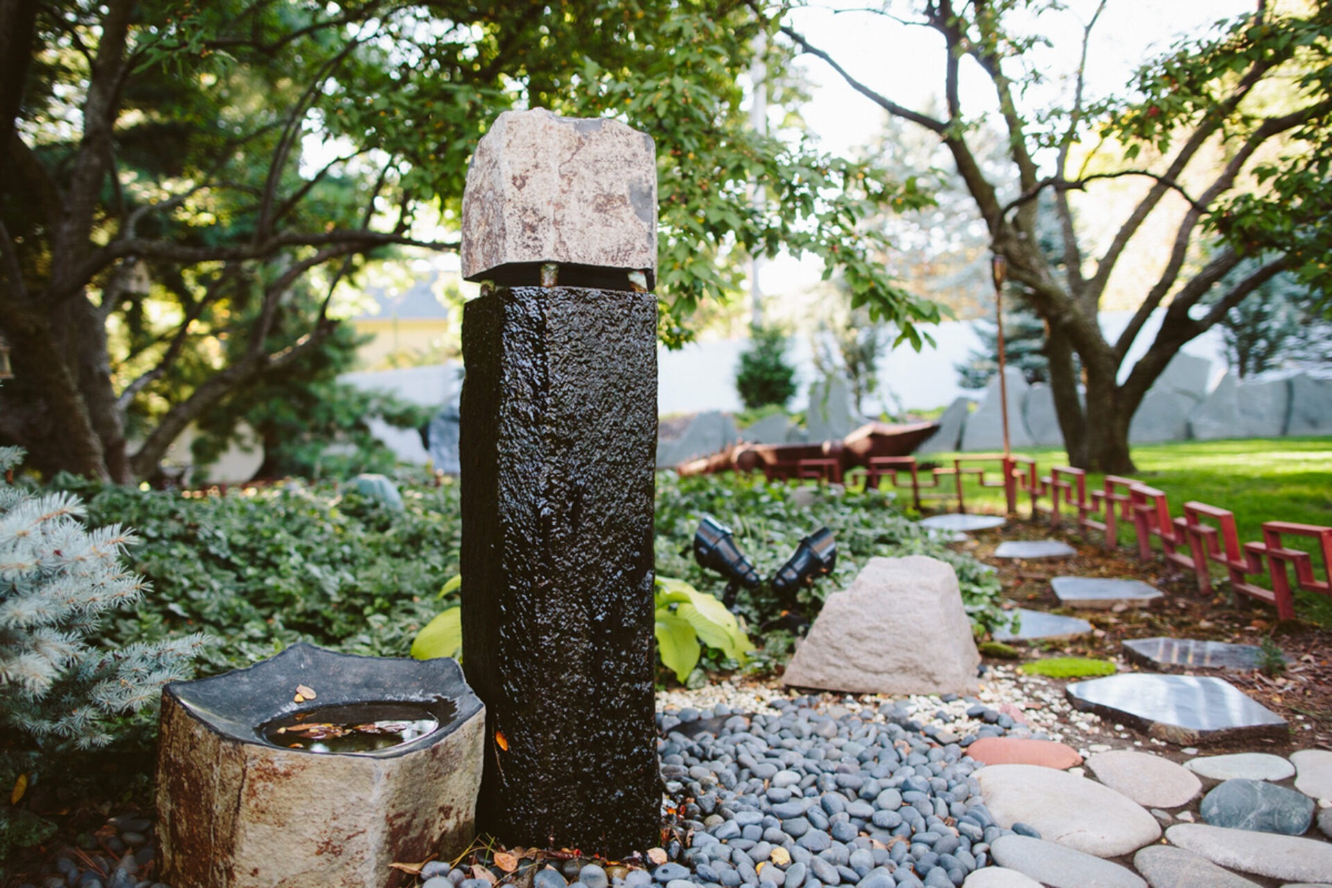 Tranquil garden scene with stone pillar, lush greenery, and a pathway of round stones under a canopy of trees, creating a peaceful atmosphere.