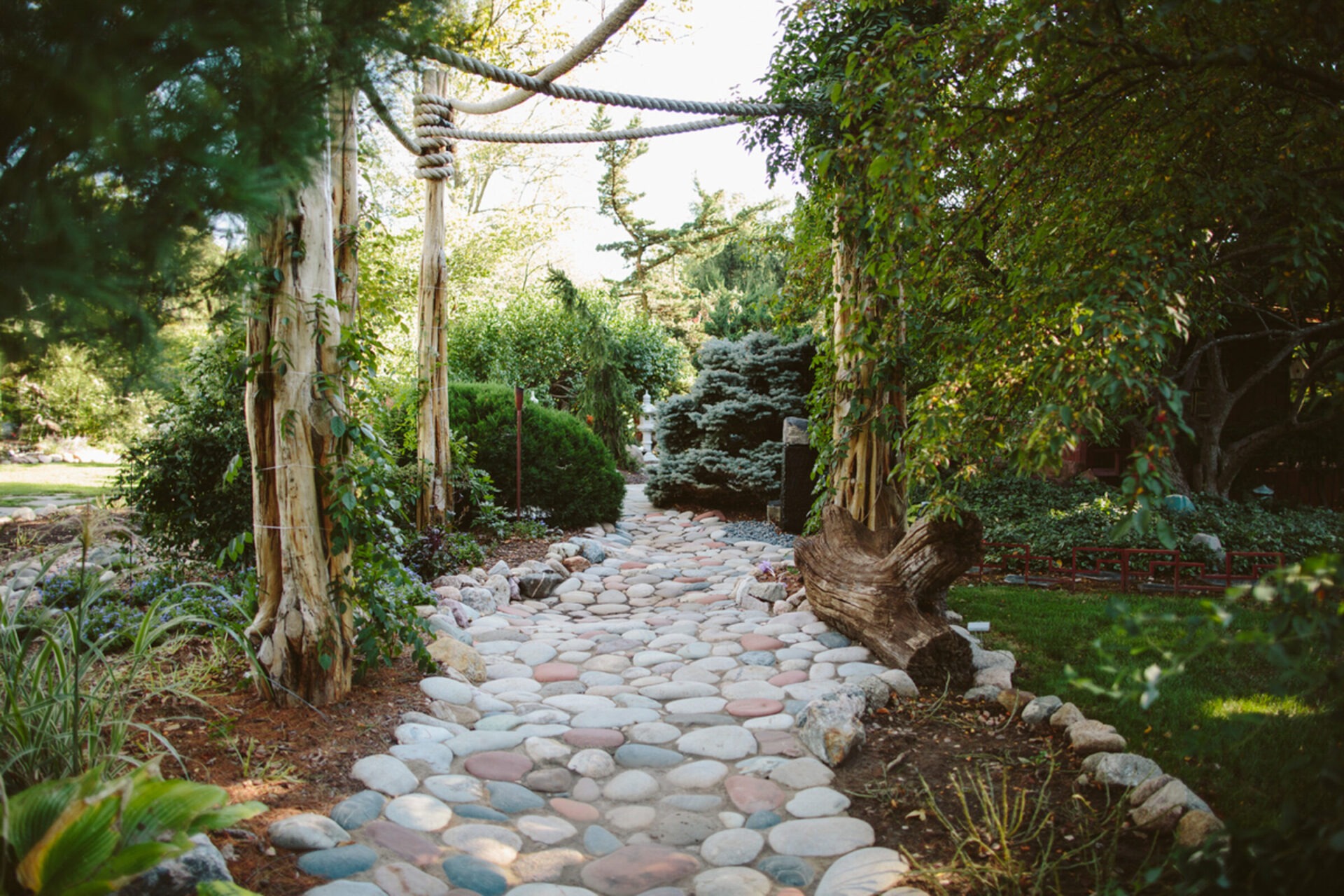 A peaceful garden scene featuring a cobblestone path, lush green plants, and rustic wooden archways under dappled sunlight.