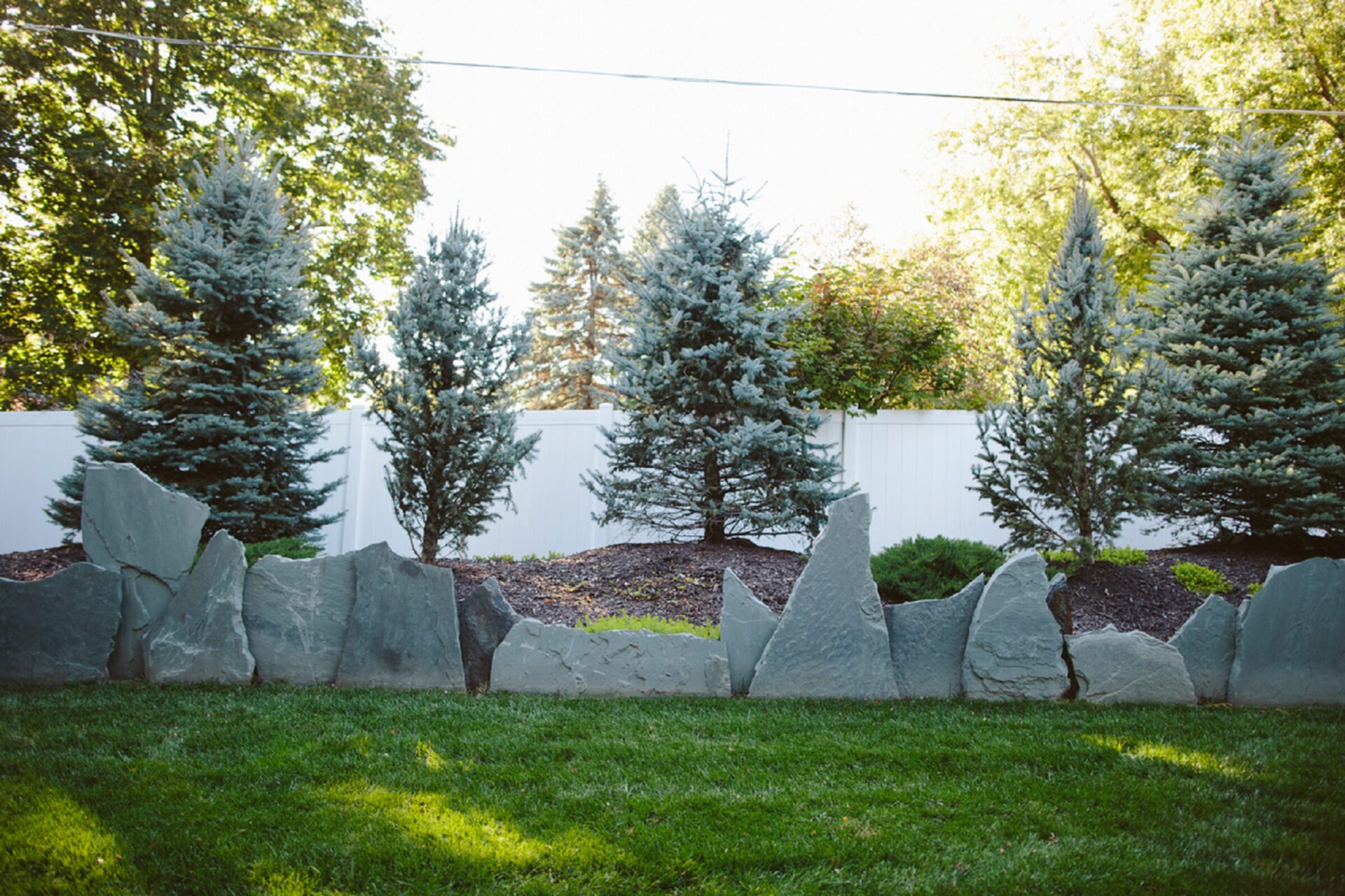 A garden scene with evergreen trees, large decorative stones, and a white fence, set against a backdrop of lush greenery and grass.