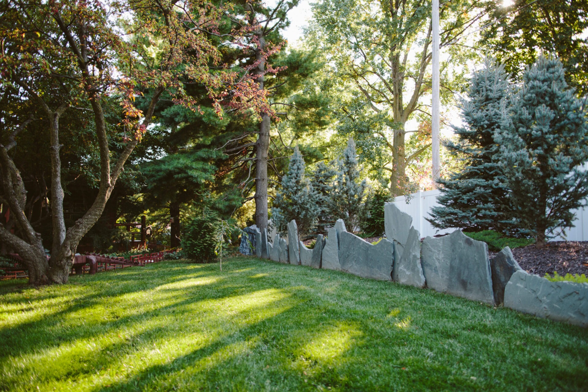 A serene garden with lush green grass, tall trees, and a decorative stone fence, basking in sunlight and shadows.