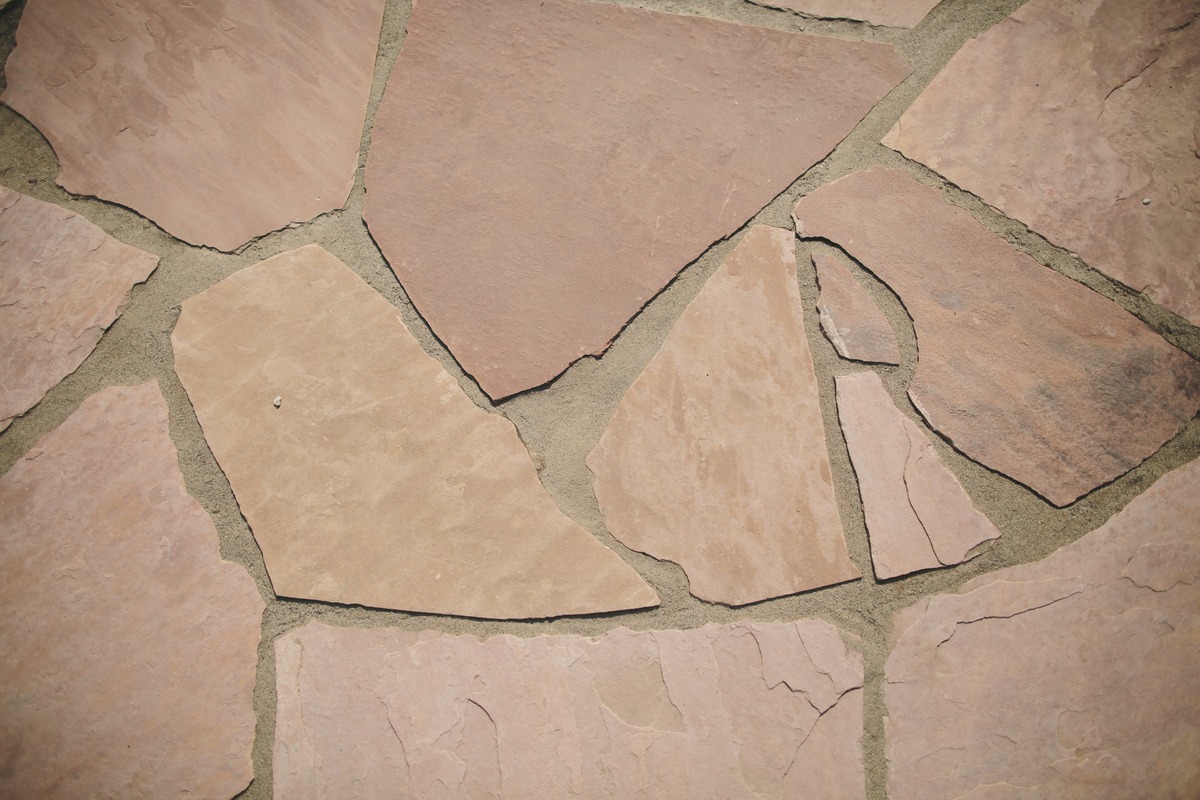 Close-up of an irregular stone pavement with various shades of beige and light brown, showcasing natural textures and gaps filled with sand.