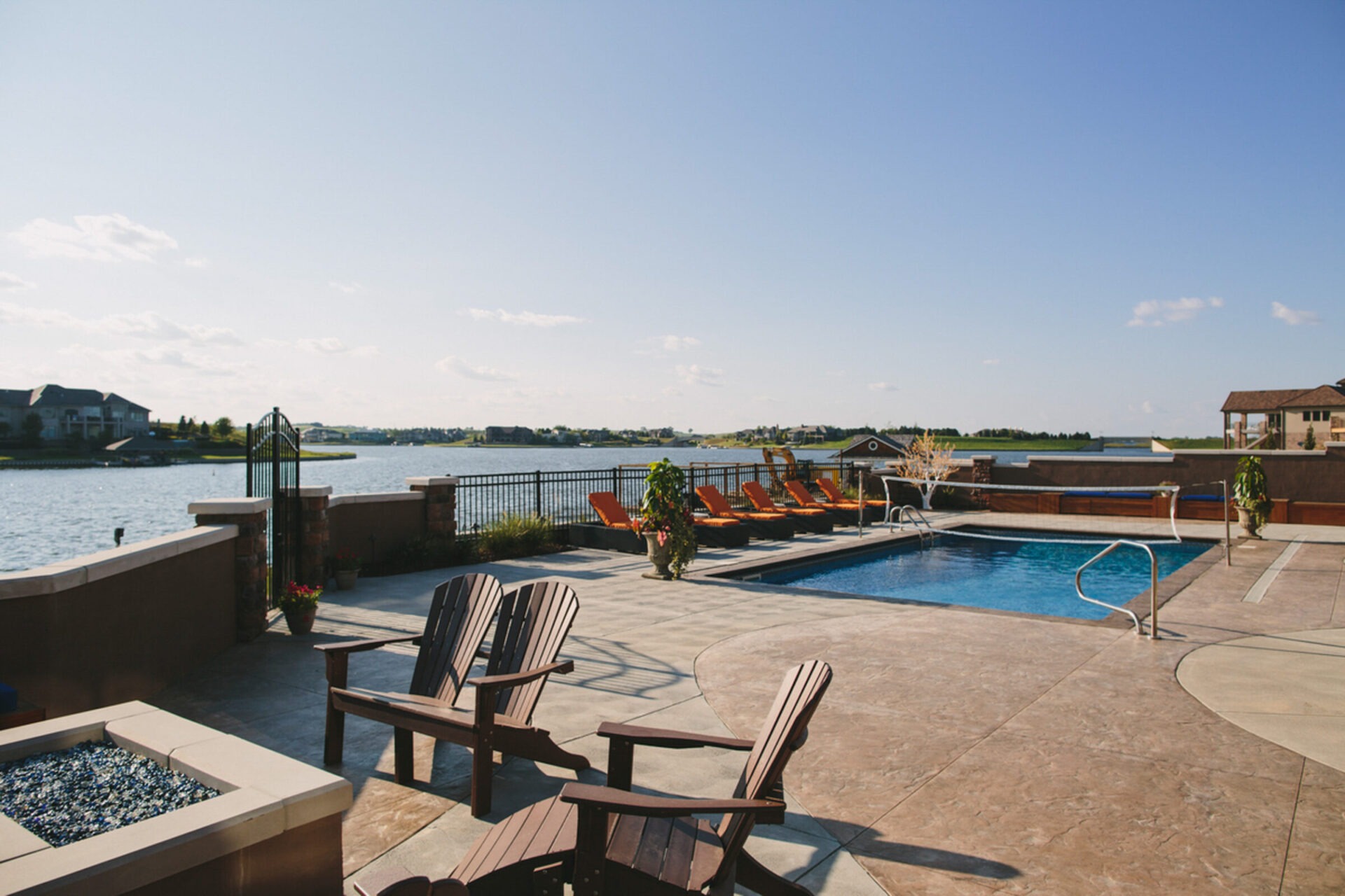 Lakeside patio with a swimming pool, lounge chairs, and Adirondack chairs overlooking the water under a clear blue sky.