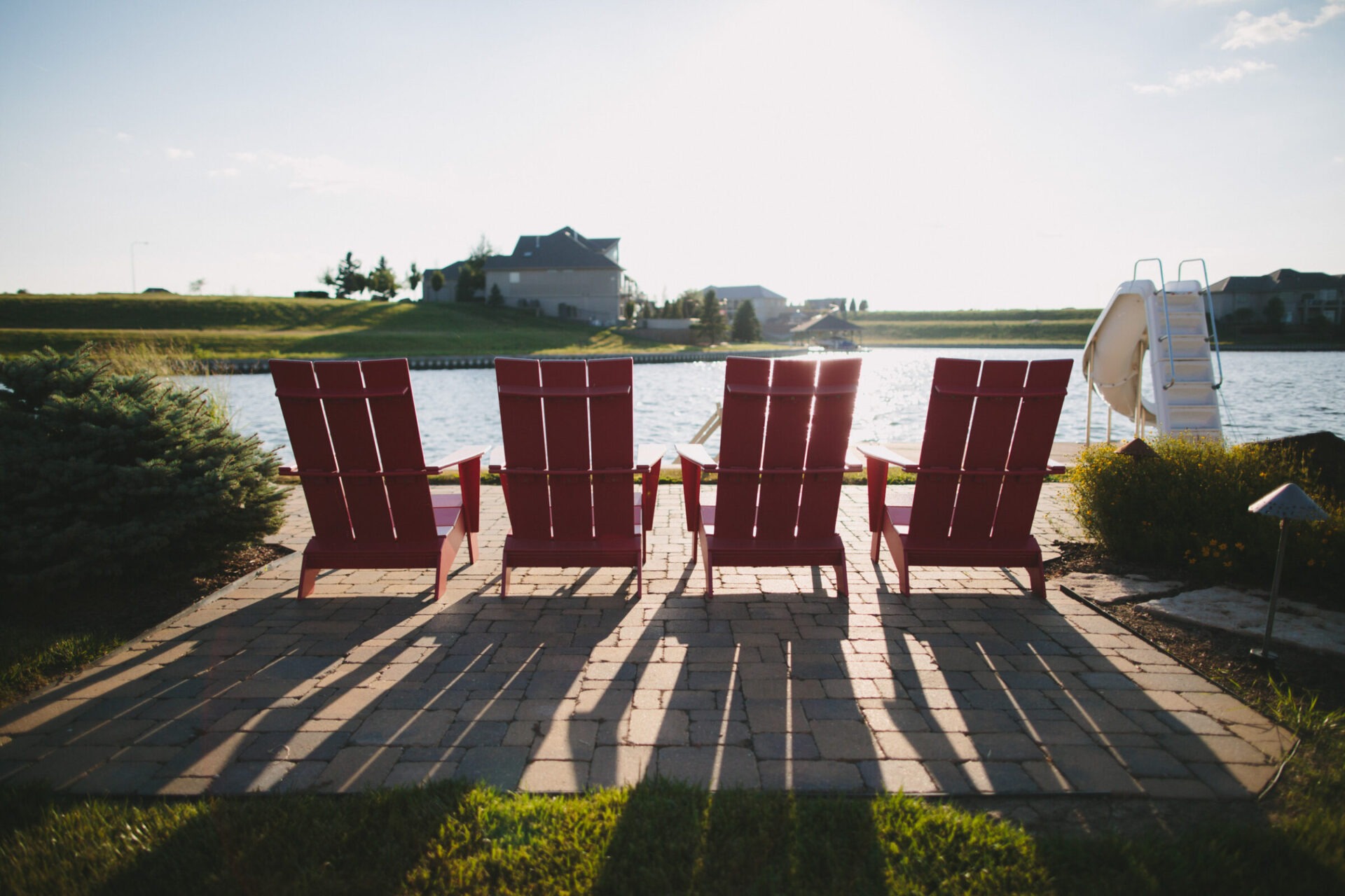 Four red chairs face a peaceful lake, surrounded by grass, shrubs, and distant houses. Sunlight casts long shadows on the patio.