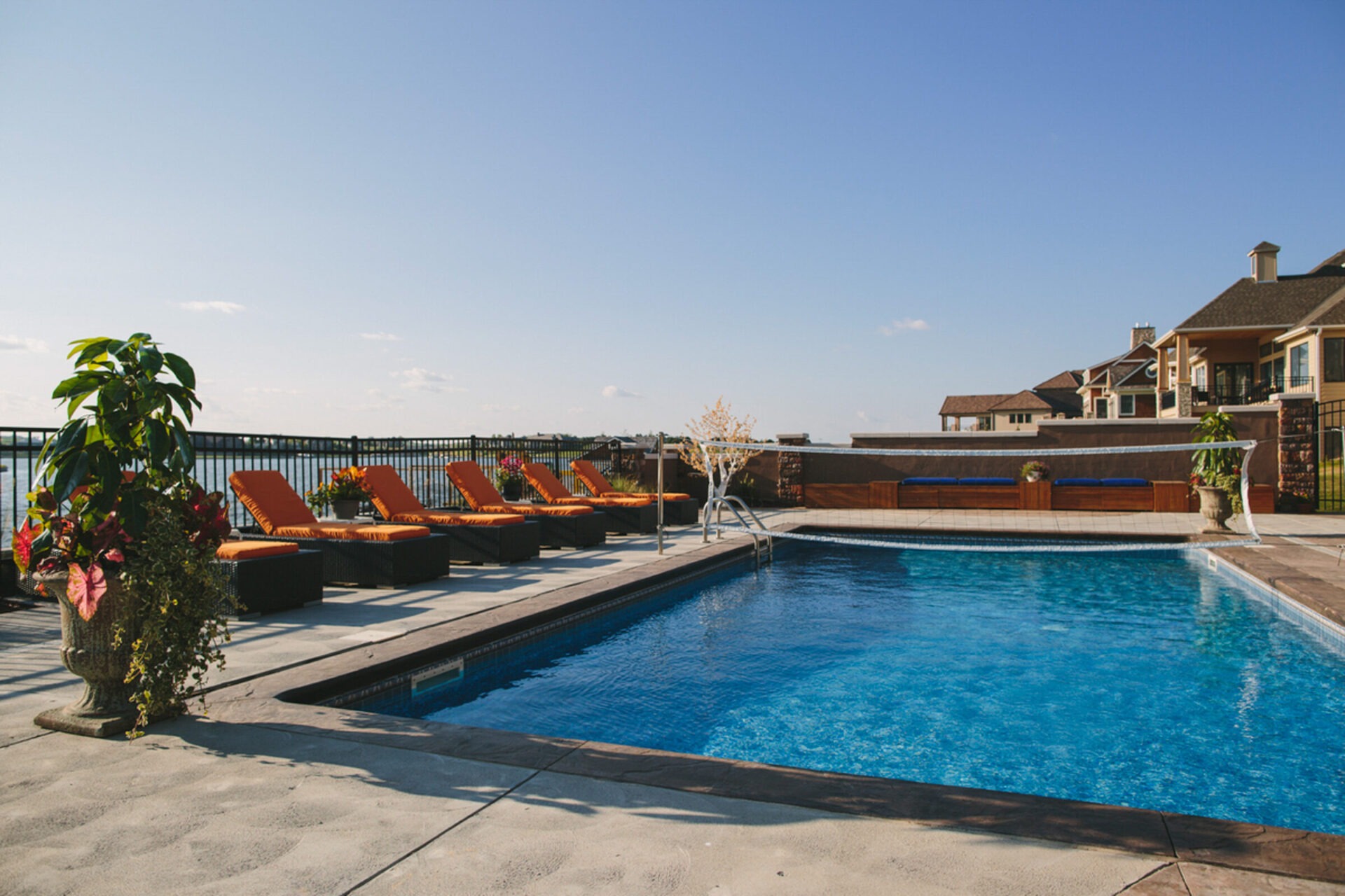 Outdoor pool with orange loungers and potted plants overlooking a water body, set against a clear sky and residential buildings.