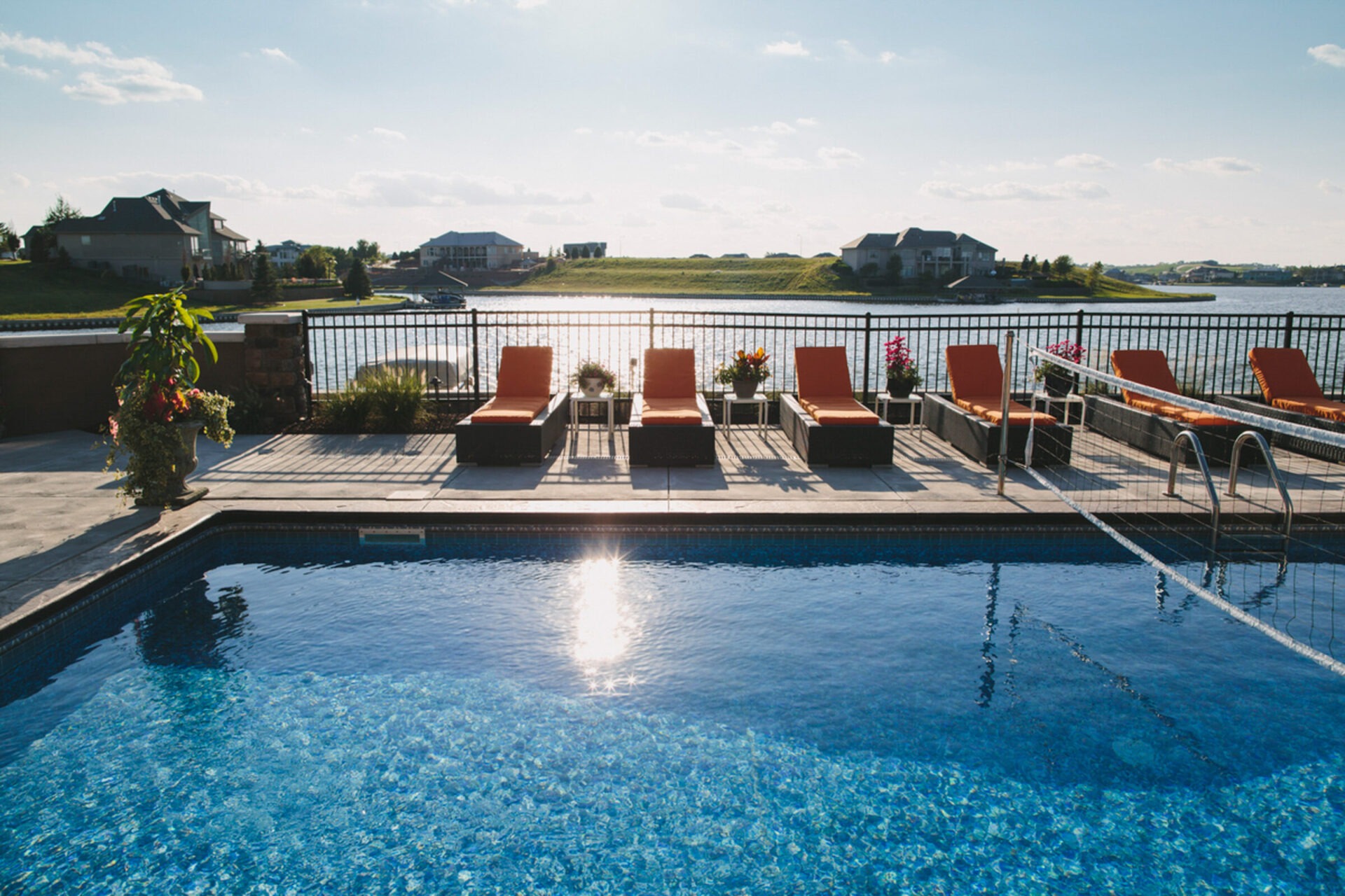 A serene poolside view with orange lounge chairs, overlooking a tranquil lake and distant suburban homes under a clear, sunny sky.