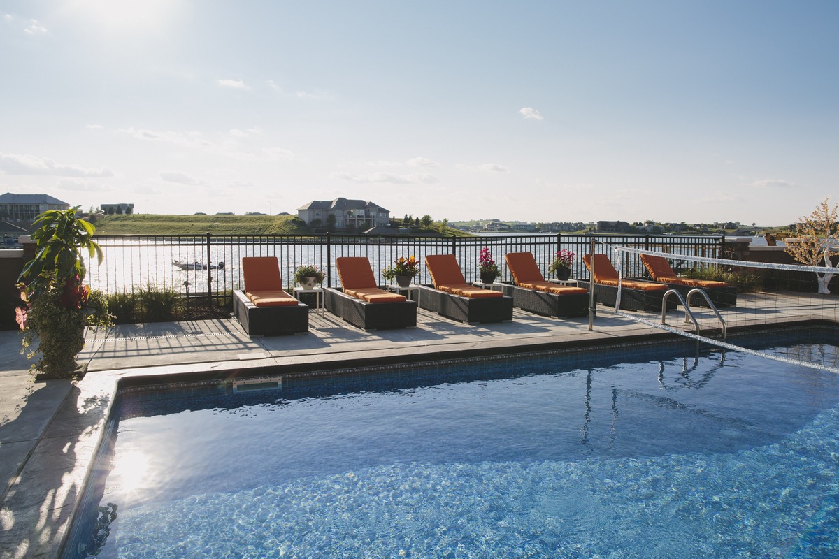 A swimming pool with lounge chairs faces a serene lake, surrounded by residential homes under a clear blue sky.