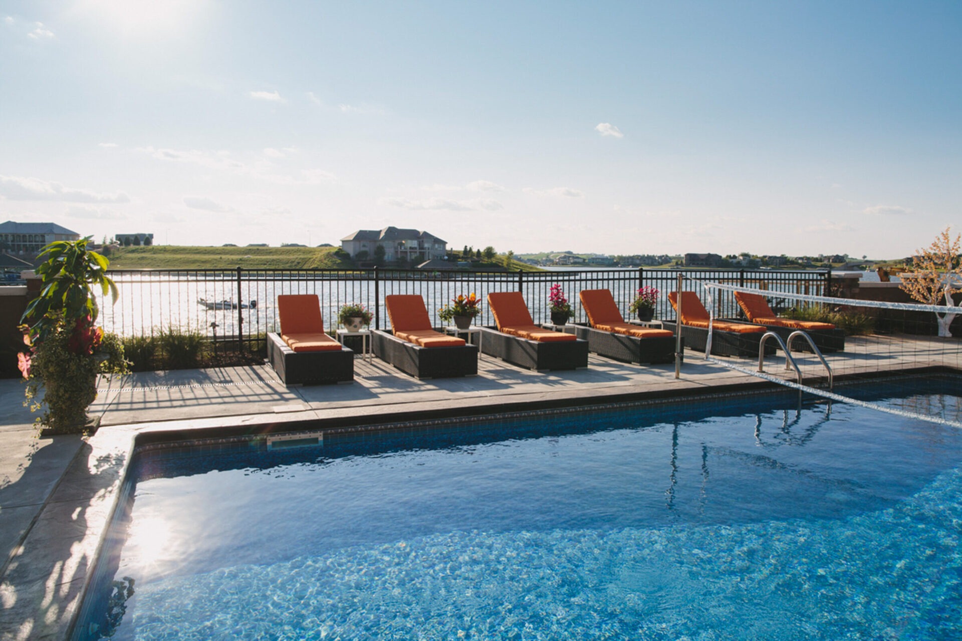 A serene outdoor pool with orange loungers overlooking a lake, surrounded by greenery and distant houses under a clear blue sky.