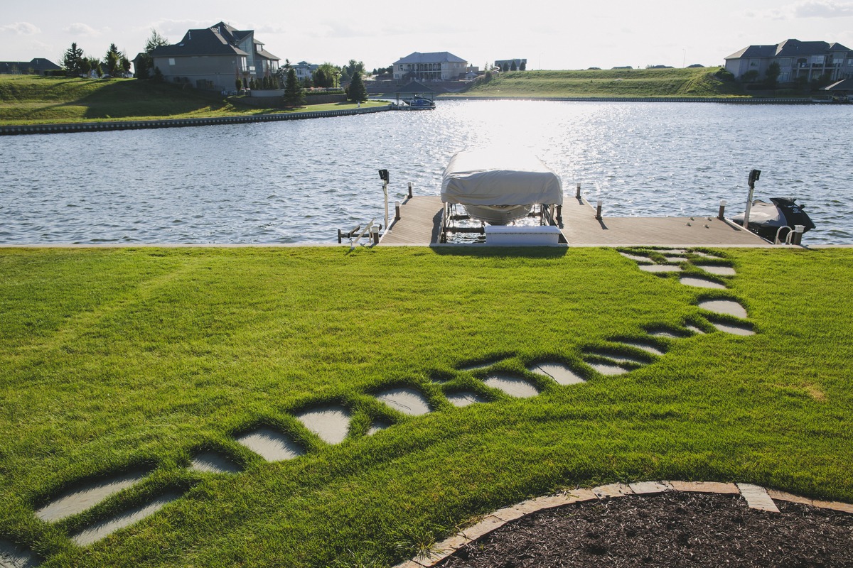 A serene lakeside scene with a docked boat, vibrant green lawn, and modern houses in the background under a bright sky.