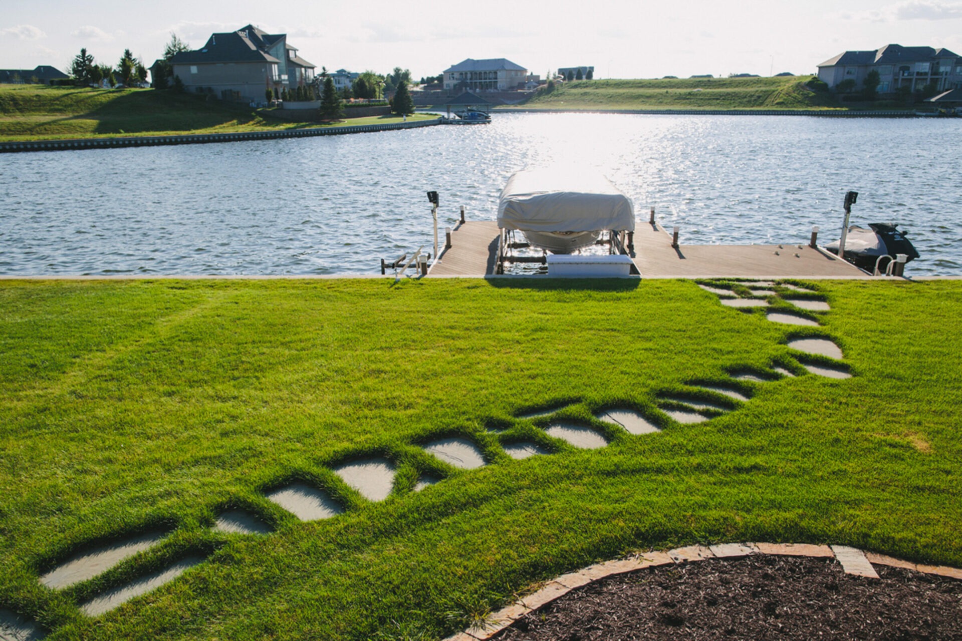 Lakeside view with a wooden dock, covered boat, and neat green lawn featuring a curved stone pathway. Houses seen in the background.