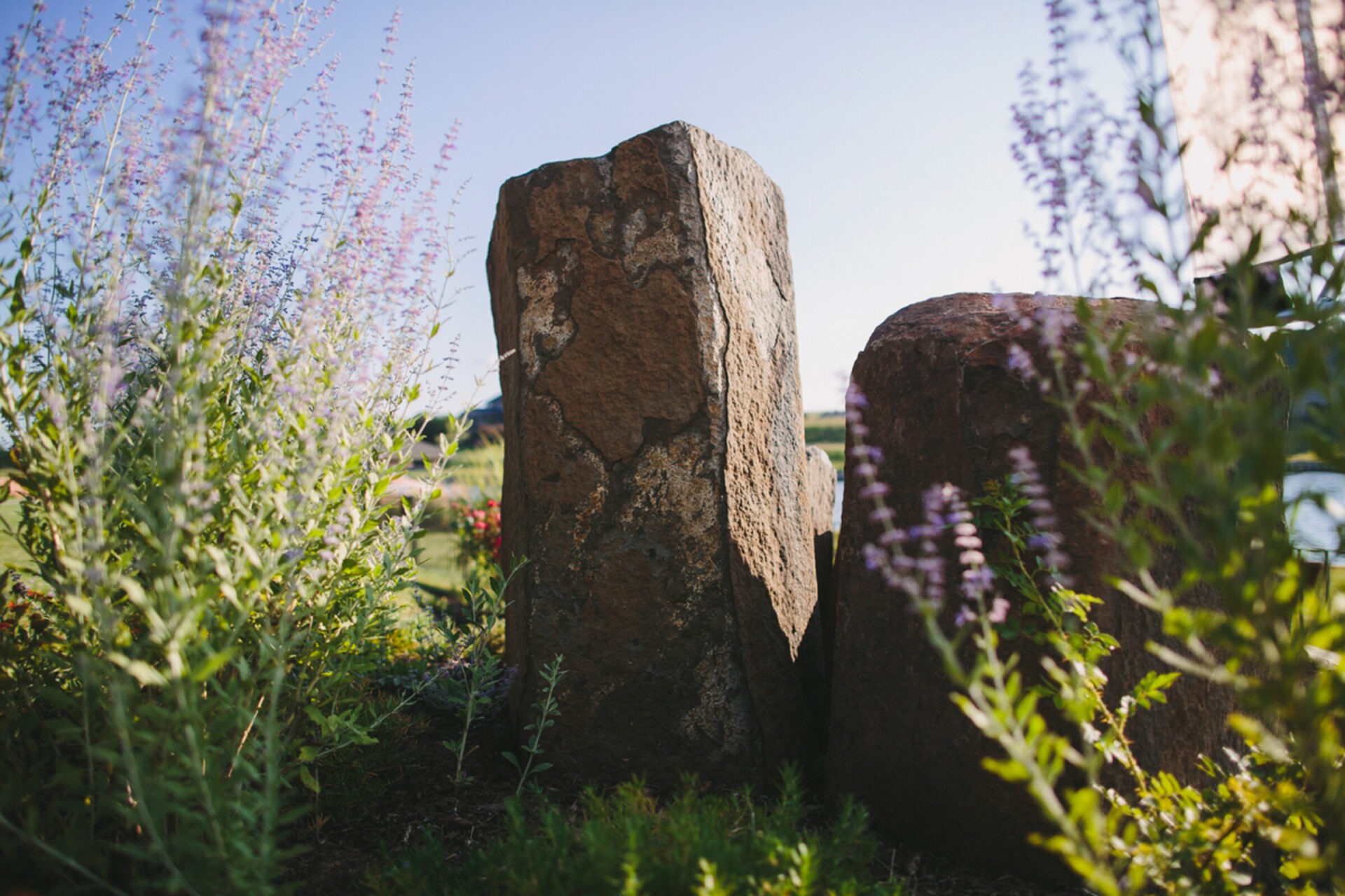 Two large stone pillars surrounded by lush greenery and purple flowers, under a clear blue sky, create a tranquil garden atmosphere.