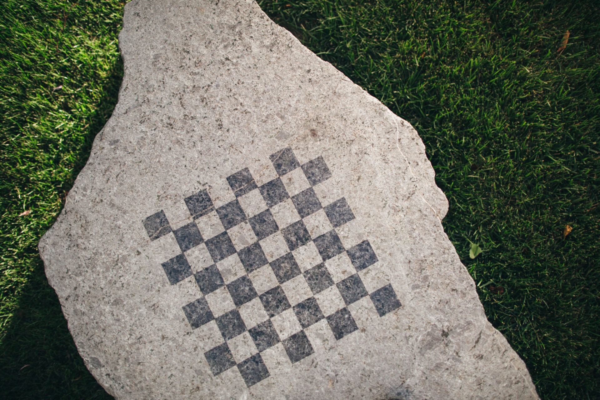 A large stone slab with a checkered pattern is set on a green grass lawn, creating a simple, outdoor design.