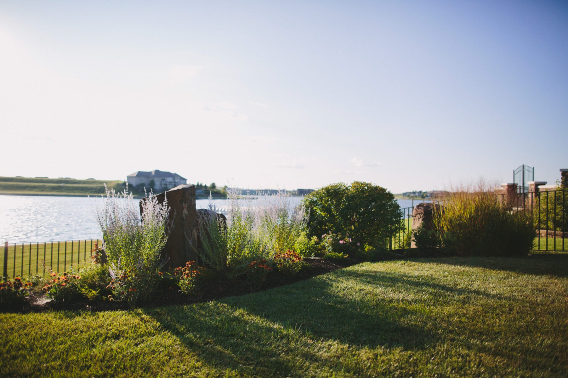 Lush garden with shrubs and flowers near a serene lake, fenced area, and distant house under a clear, sunny sky.