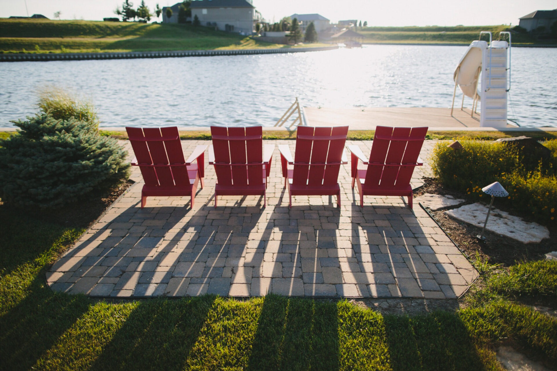 Four red chairs on a brick patio by a lake, casting long shadows, surrounded by greenery. Peaceful lakeside setting.