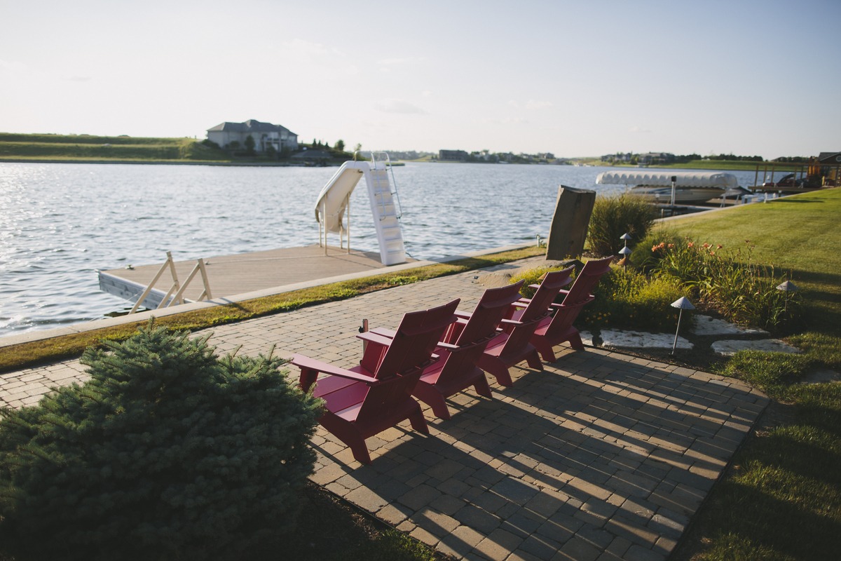 Four red chairs face a sunlit lake with a dock and slide. Greenery and a tiled walkway enhance the tranquil setting.