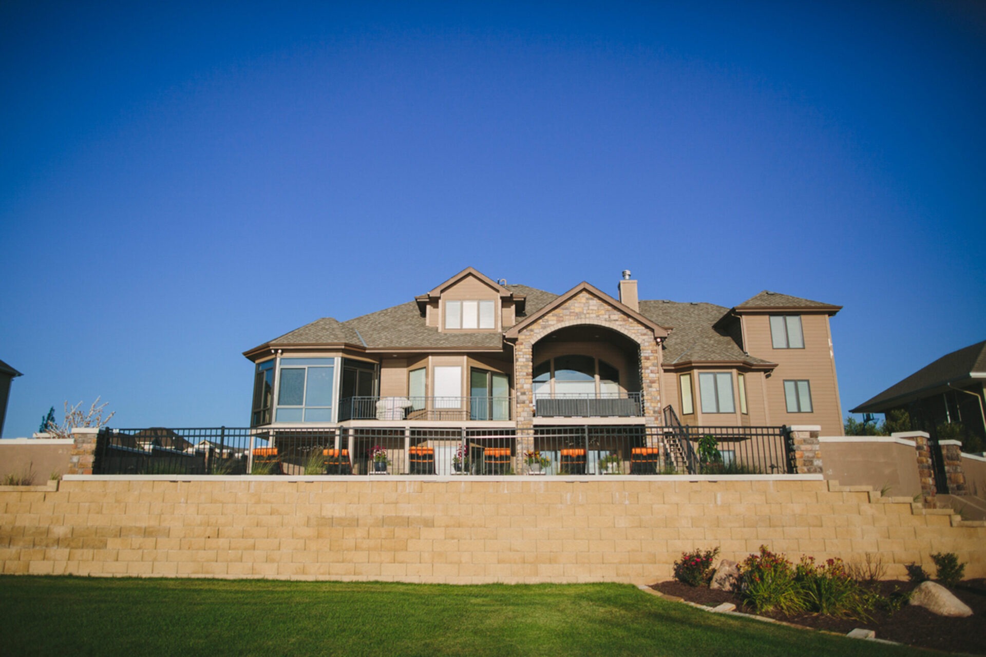 Spacious two-story house with large windows, fenced balcony, and manicured lawn under a clear blue sky. Stone entryway accentuates modern architecture.