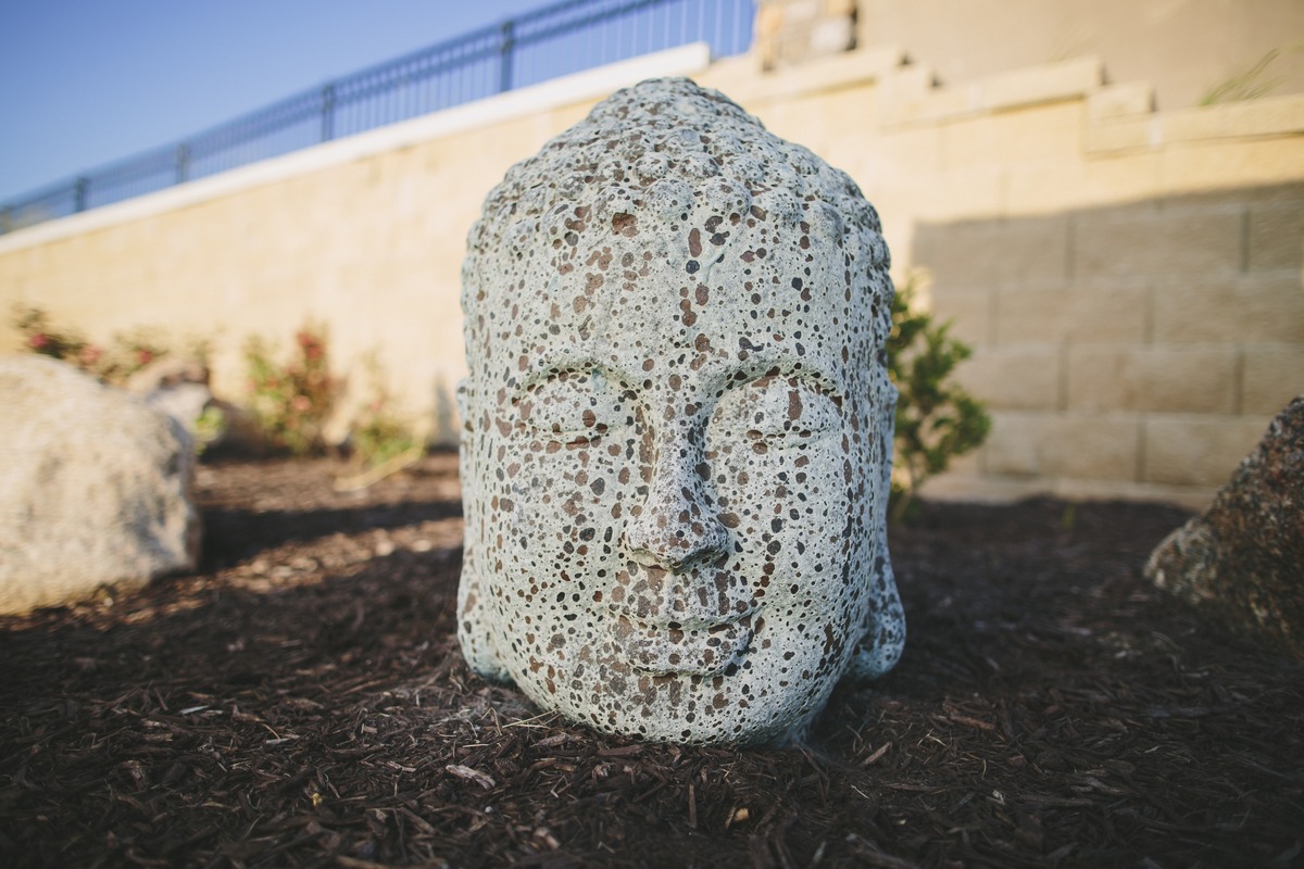 Stone sculpture of a serene face rests on mulch, surrounded by a garden. Background includes a stone wall and blue railing.