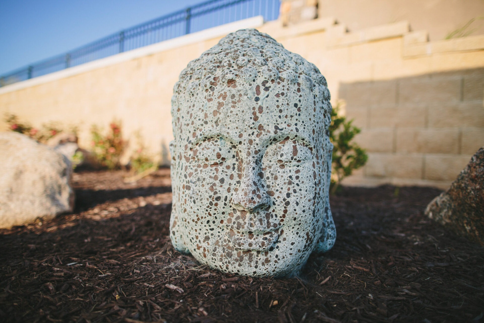 Stone sculpture of a serene face set in a garden area with mulch and plants, against a beige retaining wall and blue sky.