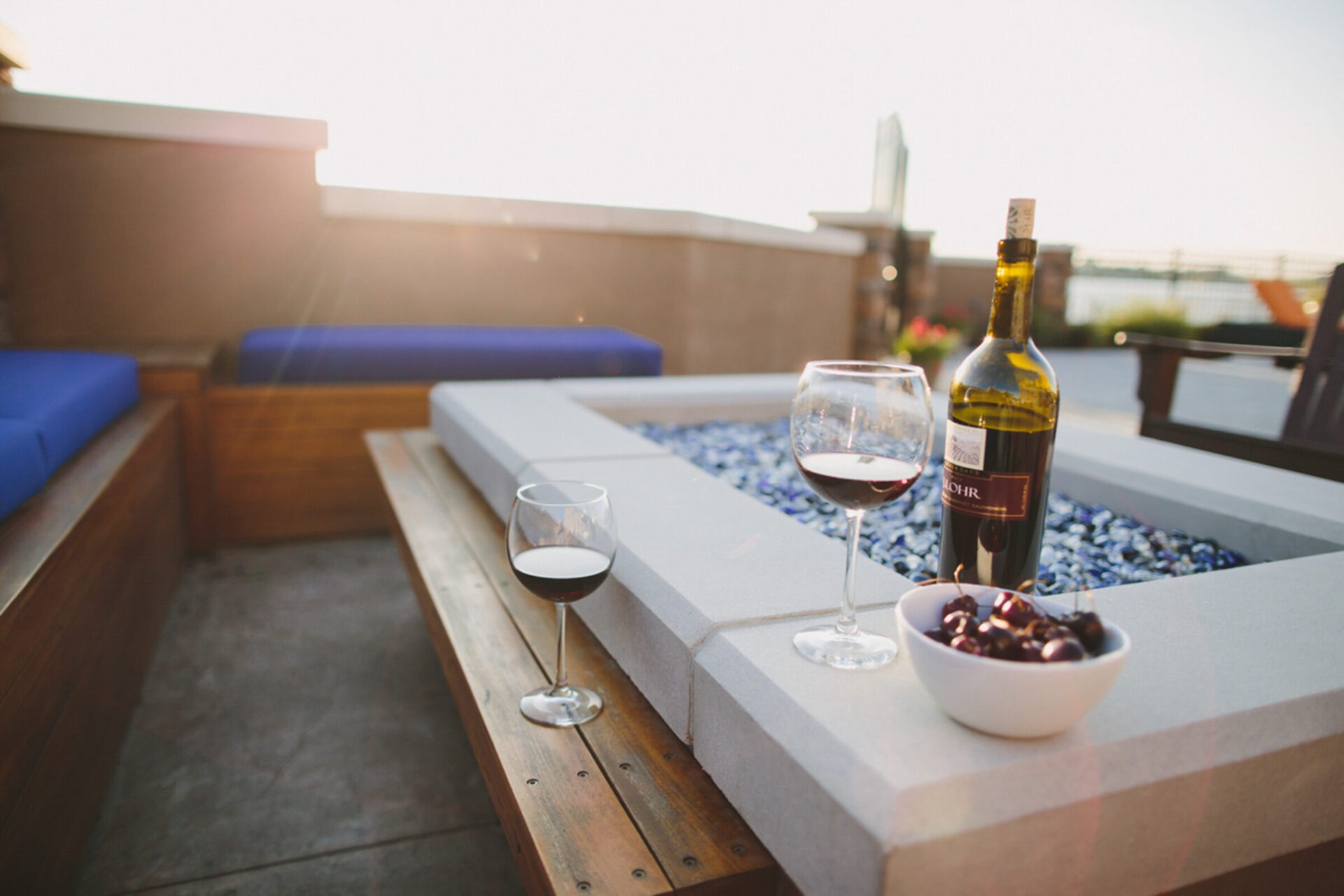 A cozy patio with a fire pit features two wine glasses, a bottle, and a bowl of cherries in natural light.