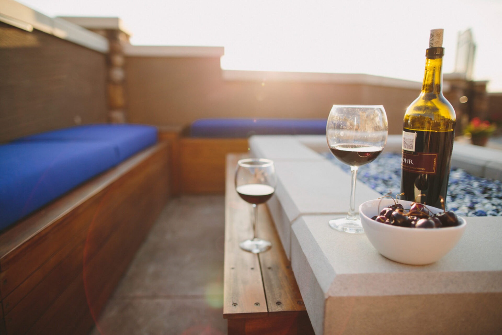 Outdoor patio setup with two wine glasses, a bottle, and a bowl of cherries on a table, featuring blue cushioned seating.