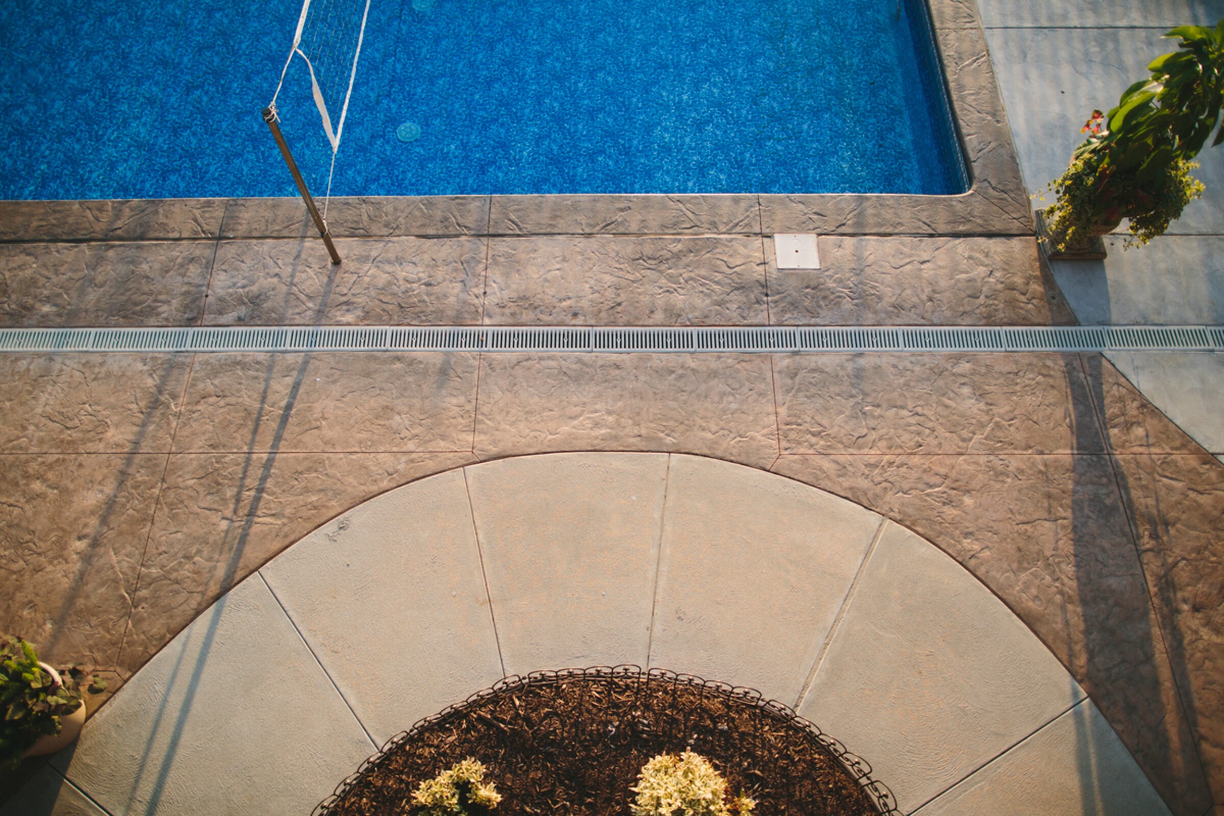 Aerial view of a swimming pool and surrounding patio with a volleyball net and potted plants; no people present.
