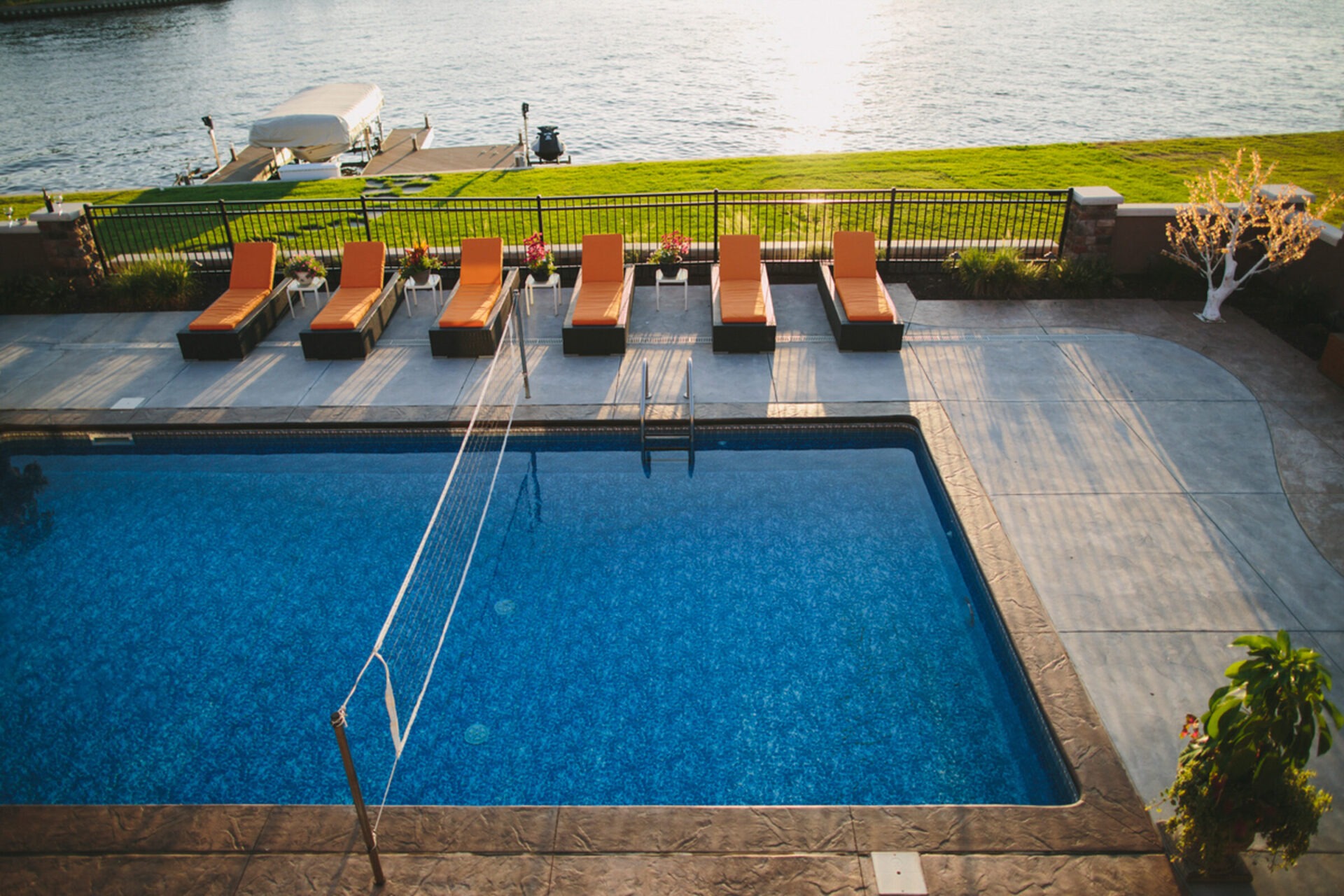 A poolside area with orange loungers overlooks a calm body of water, surrounded by lush grass and decorative plants.