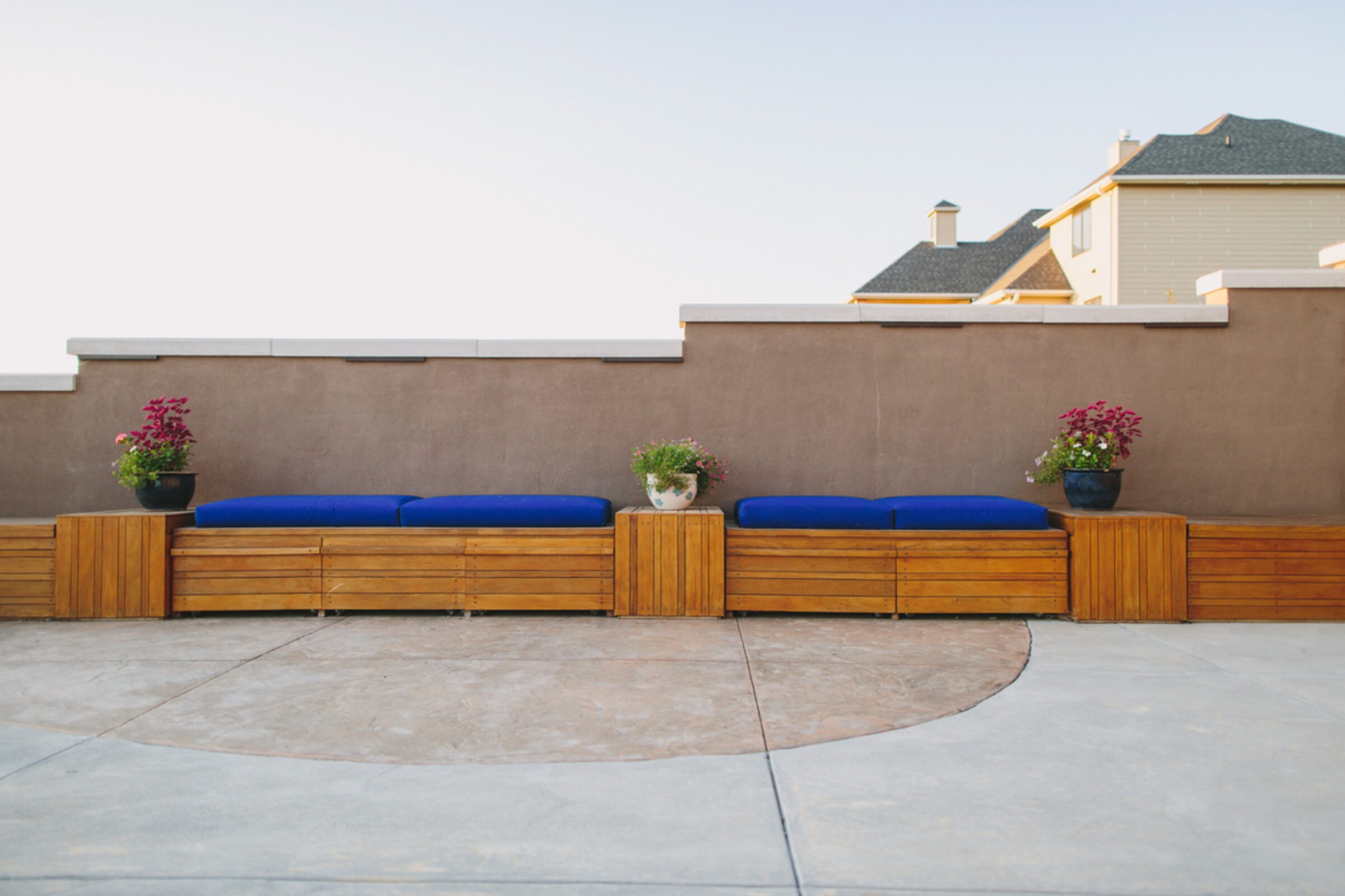 A minimalist outdoor patio with wooden benches, blue cushions, potted plants, framed by a beige wall, under a clear sky. No people present.