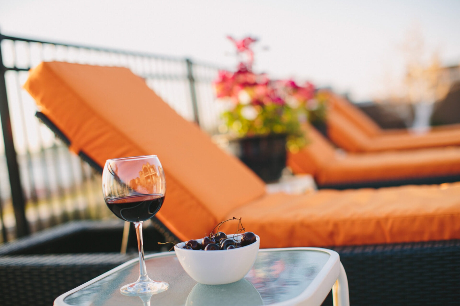 A wine glass and bowl of cherries on a table by orange lounge chairs with potted flowers in the background.