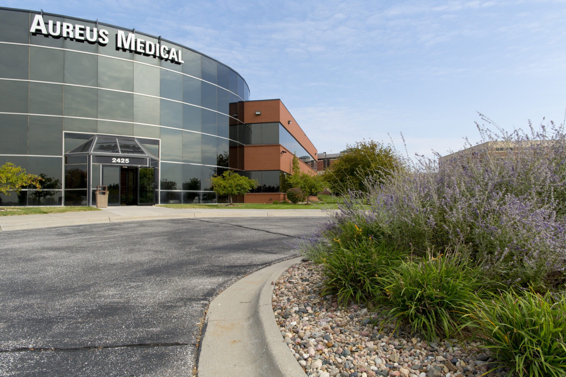 A curved glass building with "Aureus Medical" signage, surrounded by landscaped greenery and a paved driveway, under a clear sky.