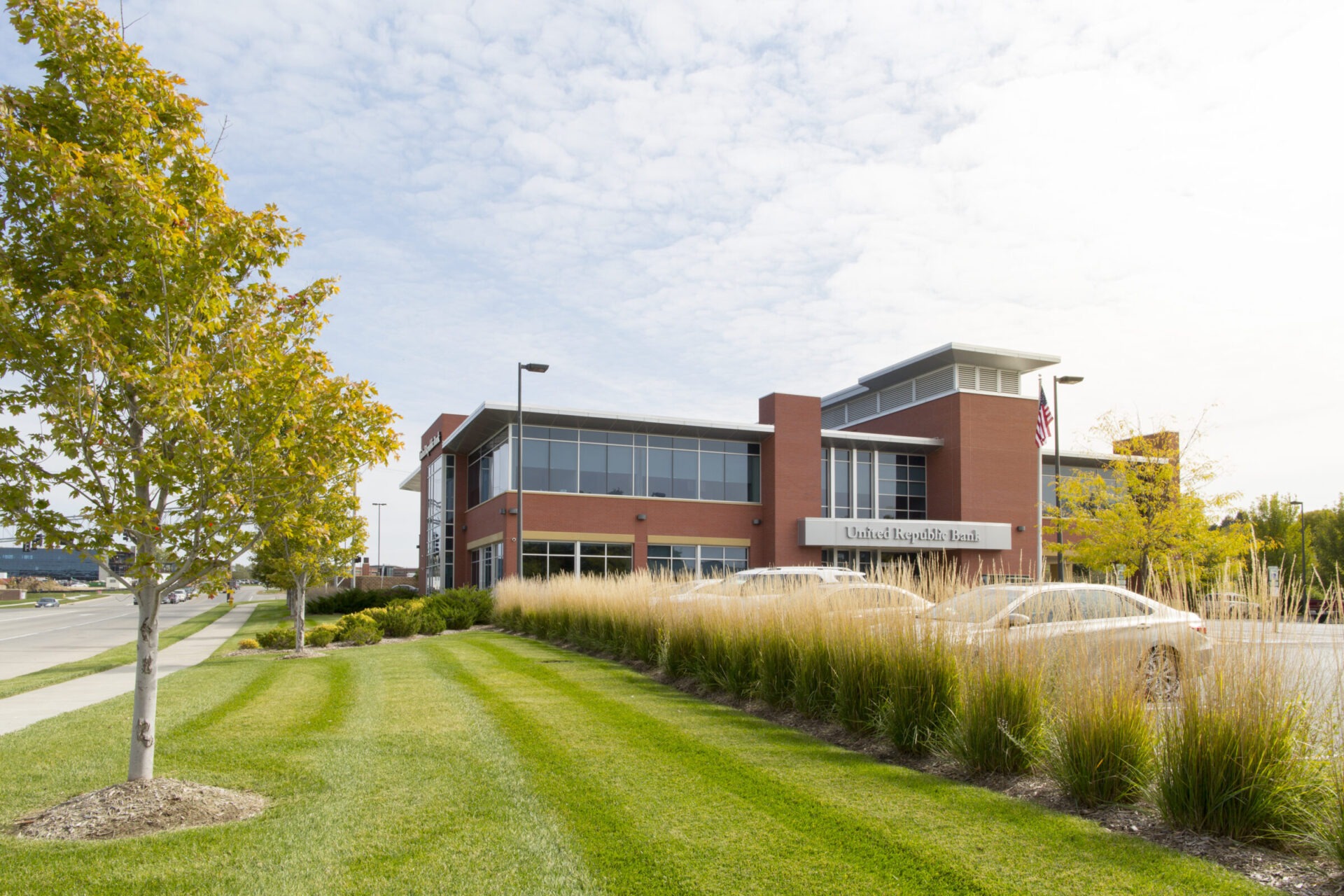 A modern brick bank building with cars parked outside, surrounded by neatly landscaped grass and trees under a partly cloudy sky.