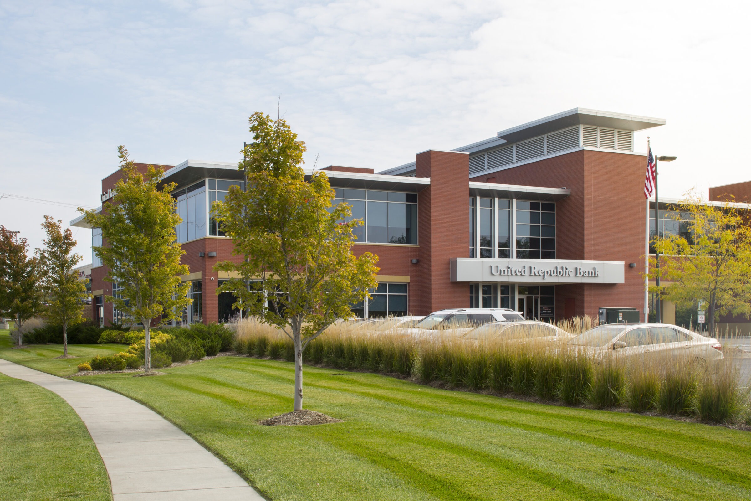 Modern brick bank building with large windows, surrounded by grass and trees. Cars are parked in front. American flag visible.