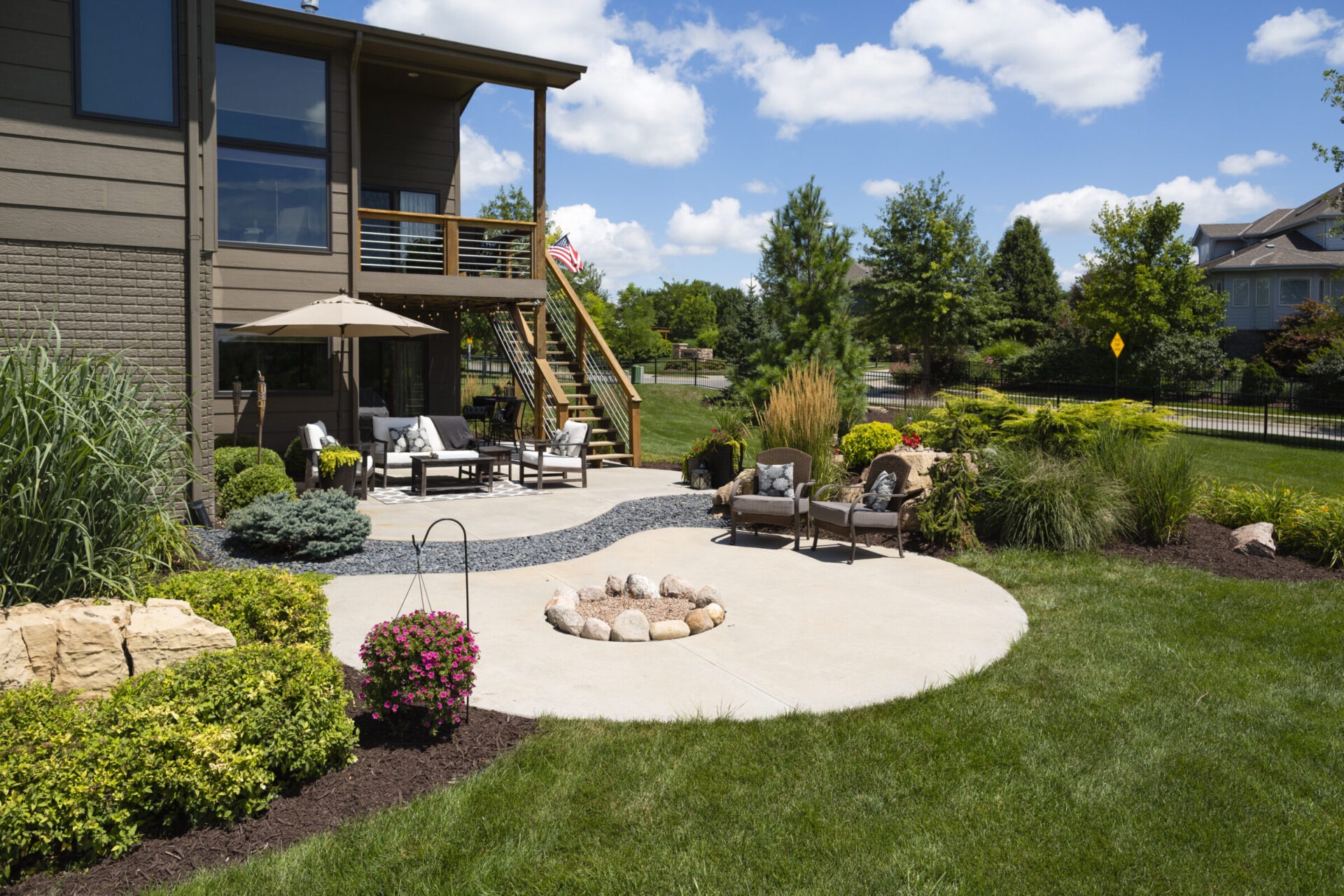 Modern backyard patio featuring seating, umbrella, and fire pit; lush landscaping with trees and bushes. Clear blue sky complements the serene setting.