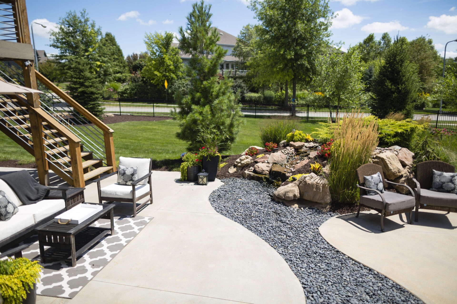 A well-designed backyard patio with comfortable seating, decorative stones, plants, and a small waterfall, set against a suburban neighborhood backdrop.