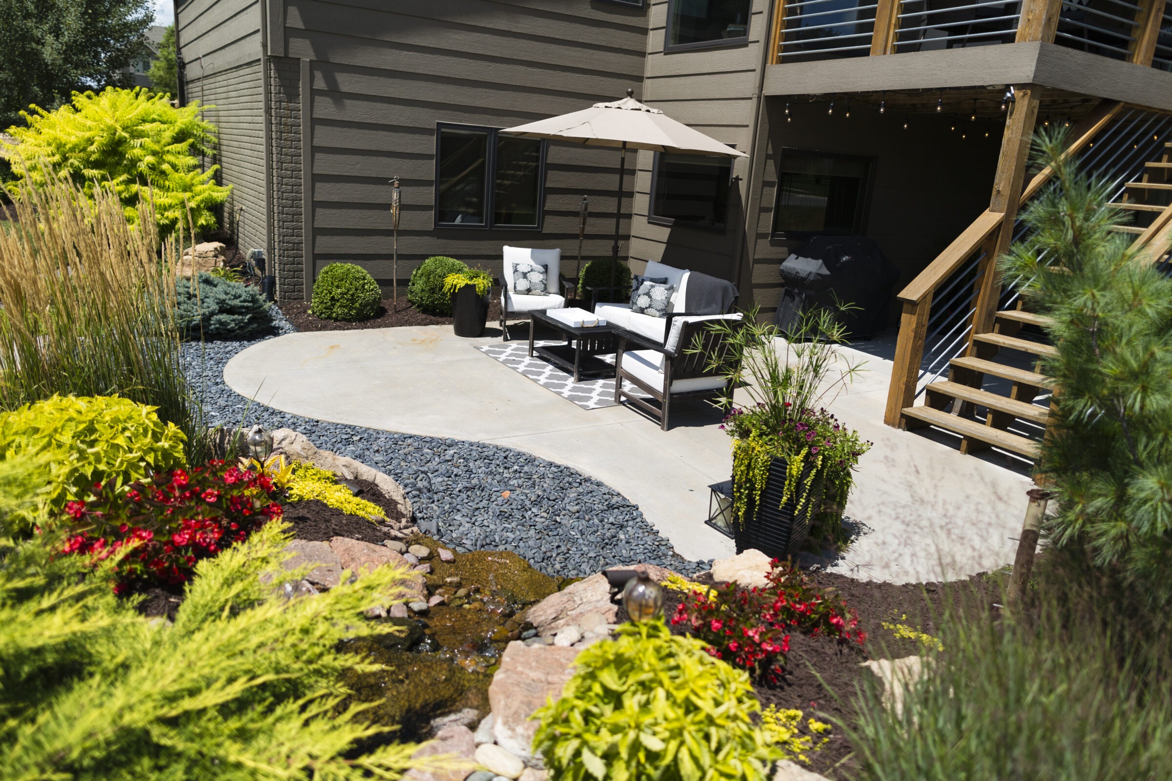 A cozy garden patio with chairs, umbrella, and decorative plants. Steps lead to a wooden deck. No people or landmarks visible.