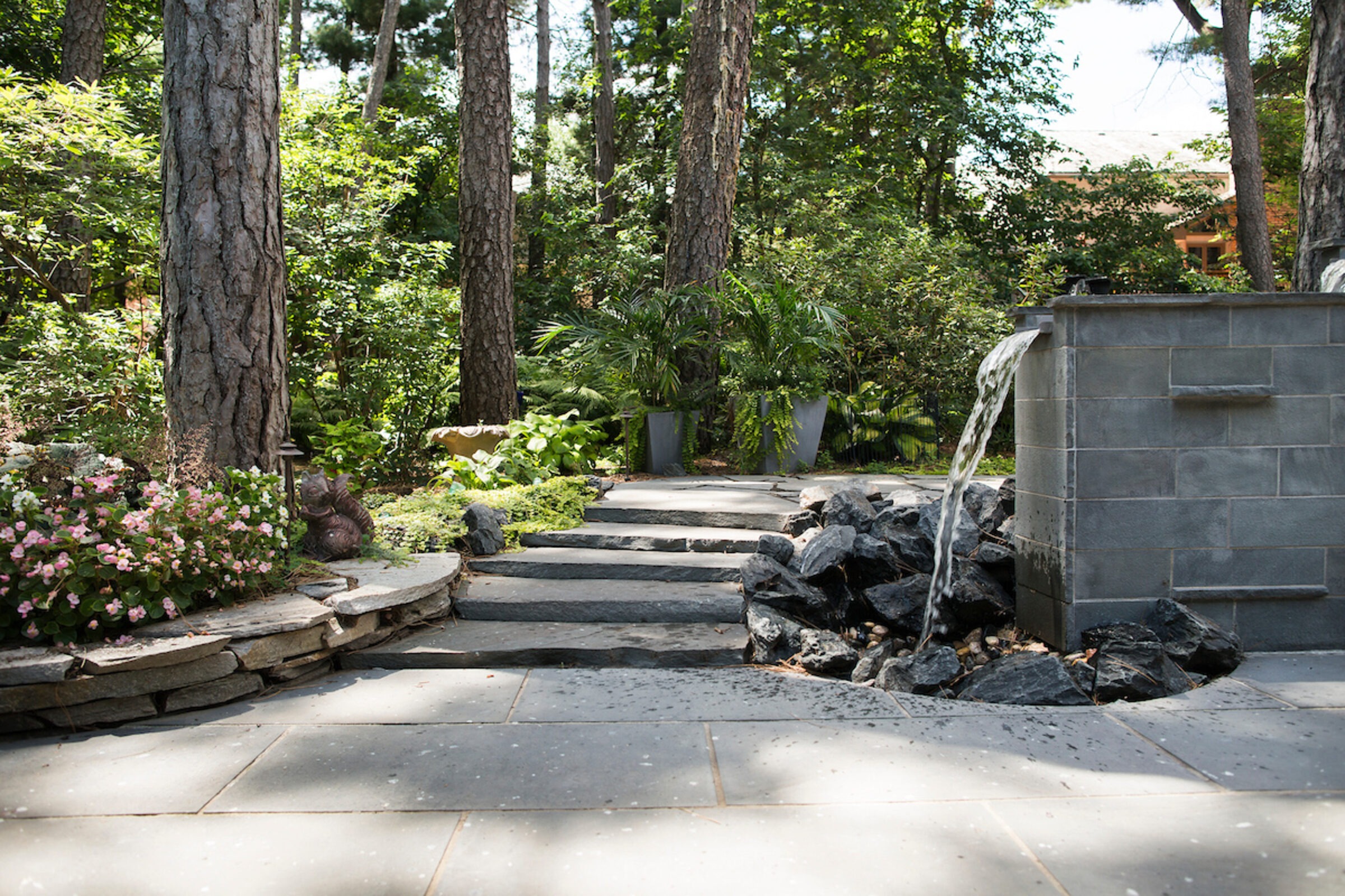 A serene garden with a stone path, abundant greenery, a water feature, and pink flowers, surrounded by tall trees under sunlight.